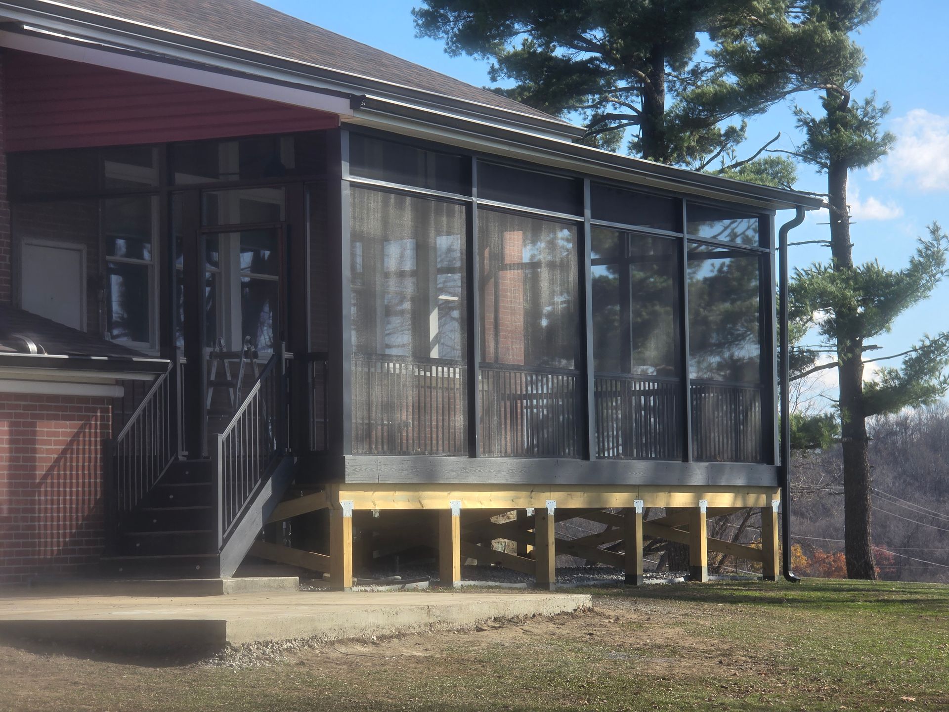 Screened-in porch with wooden deck and black frame. Red brick building and tall pine tree in the background.
