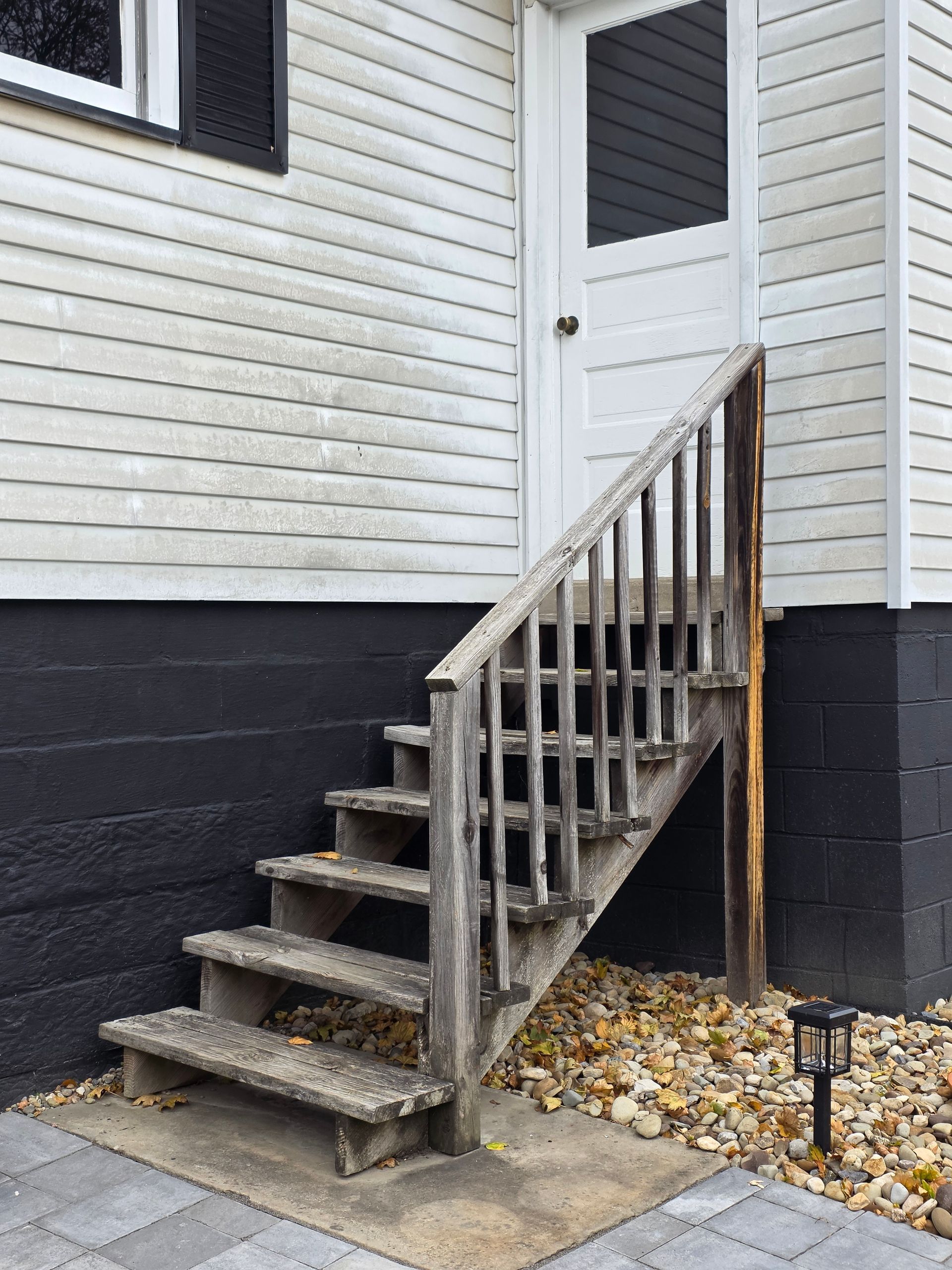 Wooden stairs leading to a white door on a building with white siding and black painted foundation.