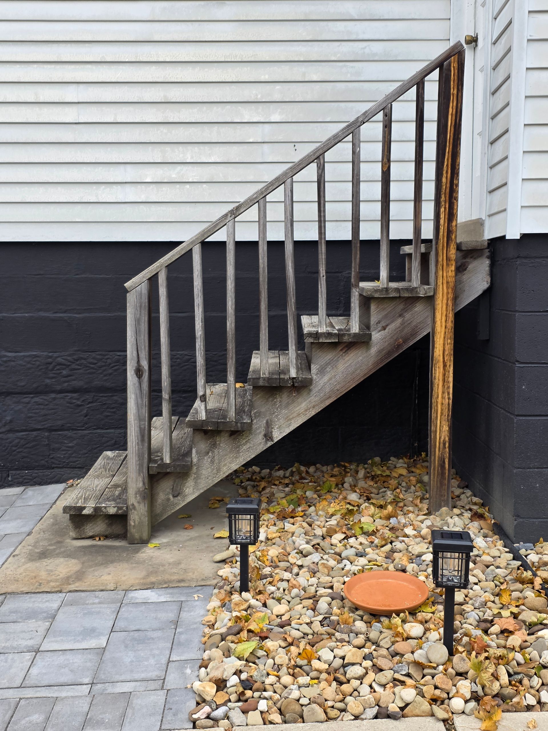 Outdoor wooden stairs next to a building with black and white walls. Two solar lights and gravel are at the base.