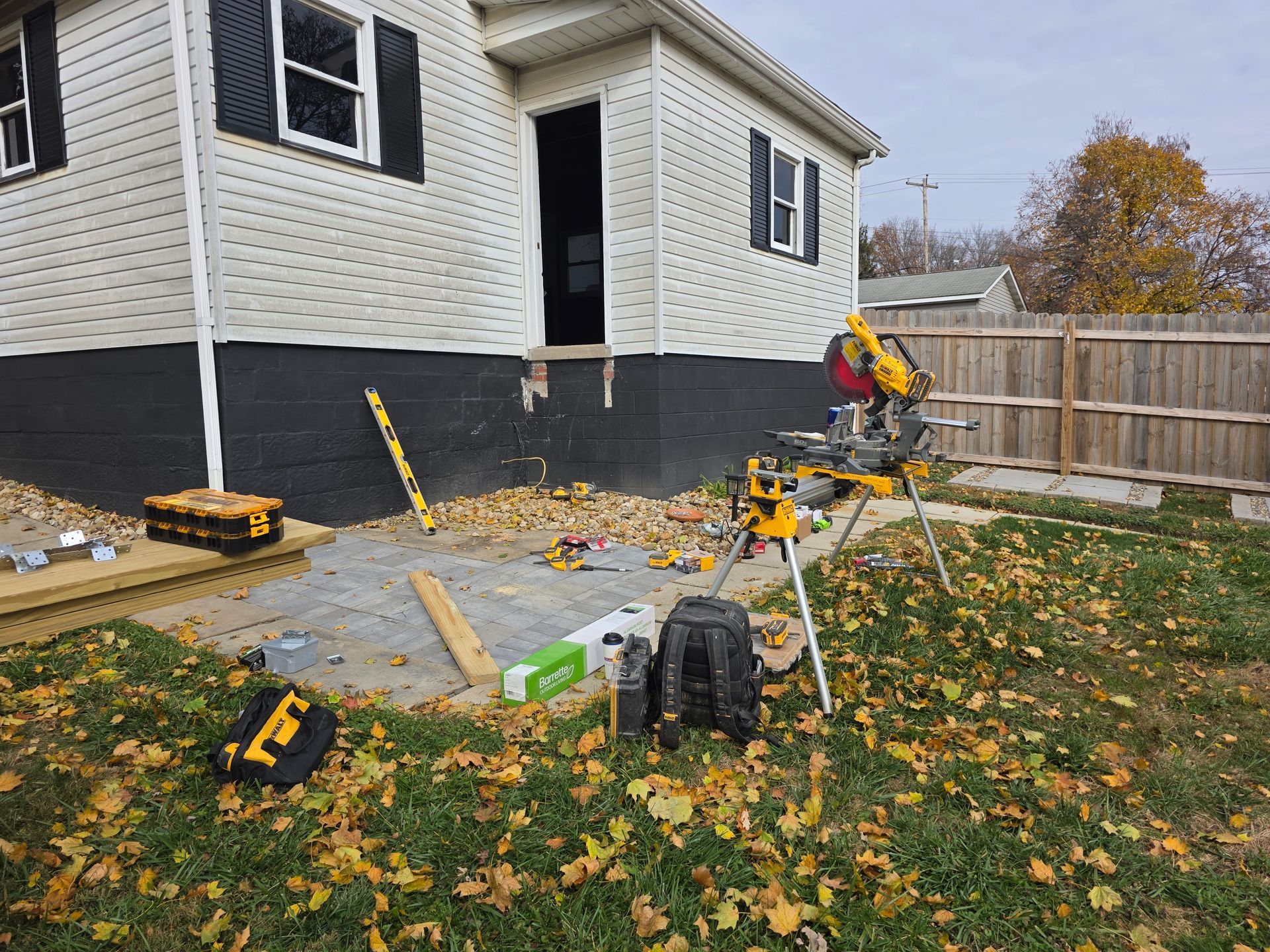 Exterior house with open doorway, tools, and construction materials scattered on a concrete patio covered in autumn leaves.