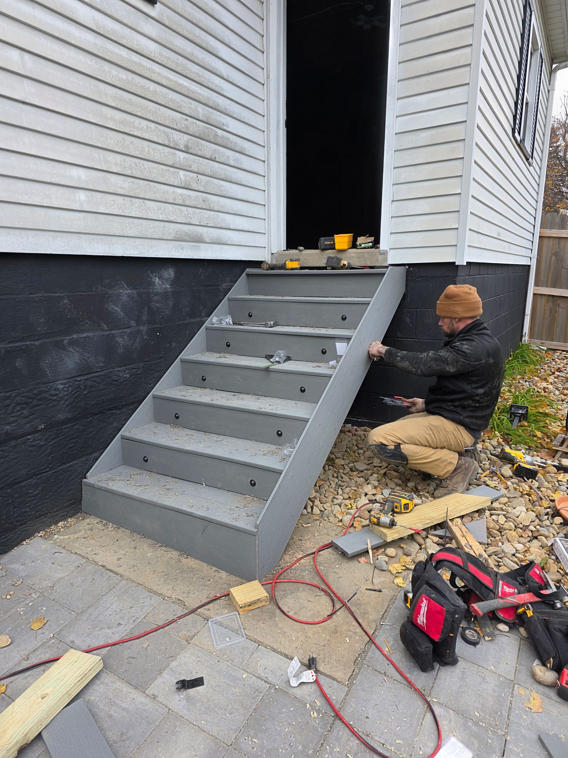 Person installing side trim on gray concrete steps leading to a doorway. Outdoor setting, tools visible.