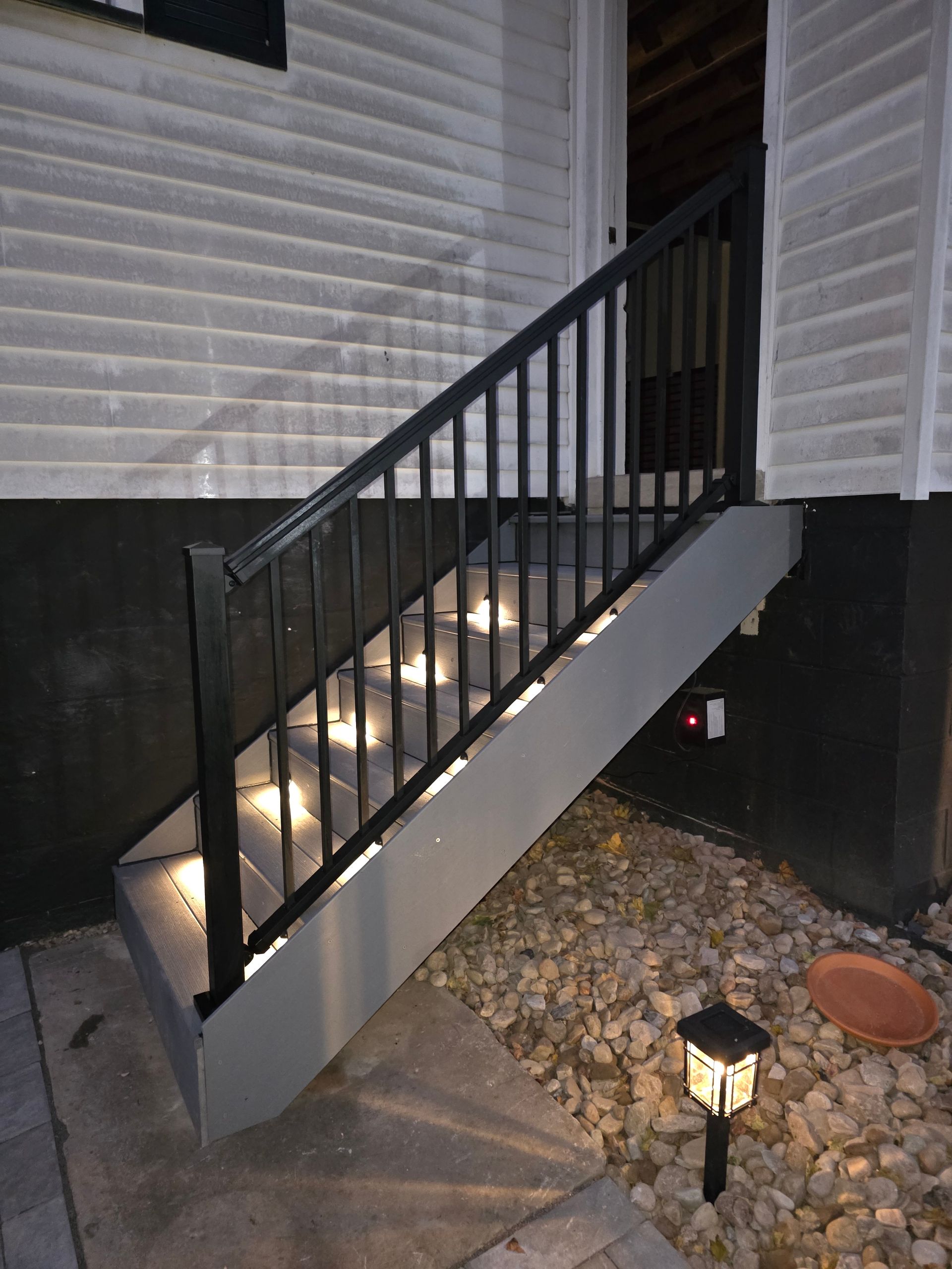 Outdoor staircase with built-in lights, black railing, leading up to a white building. Ground level has rock and a pathway.