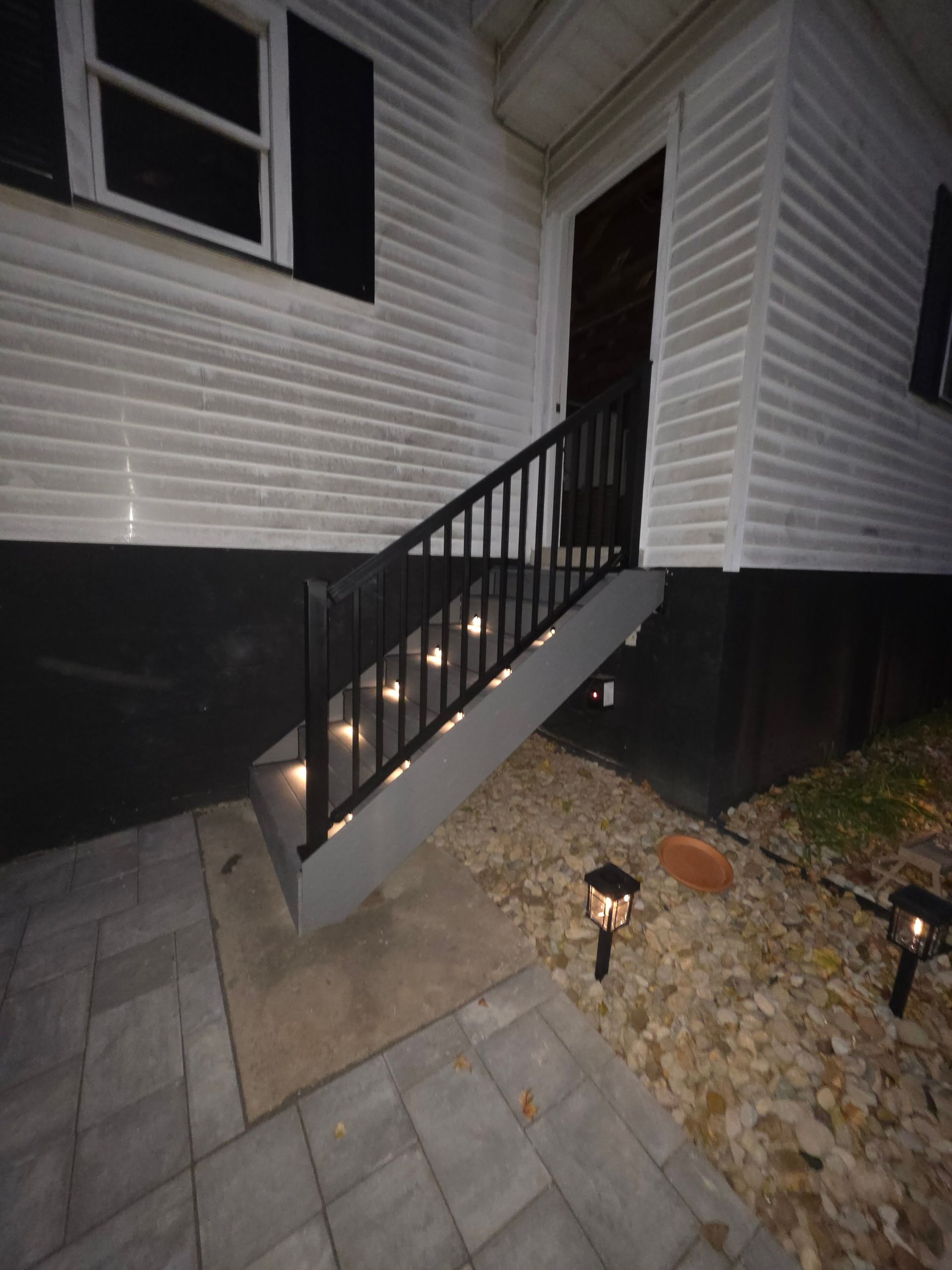 Exterior view of a house's entrance with steps illuminated by small lights, black railing. Paver walkway and garden lights.
