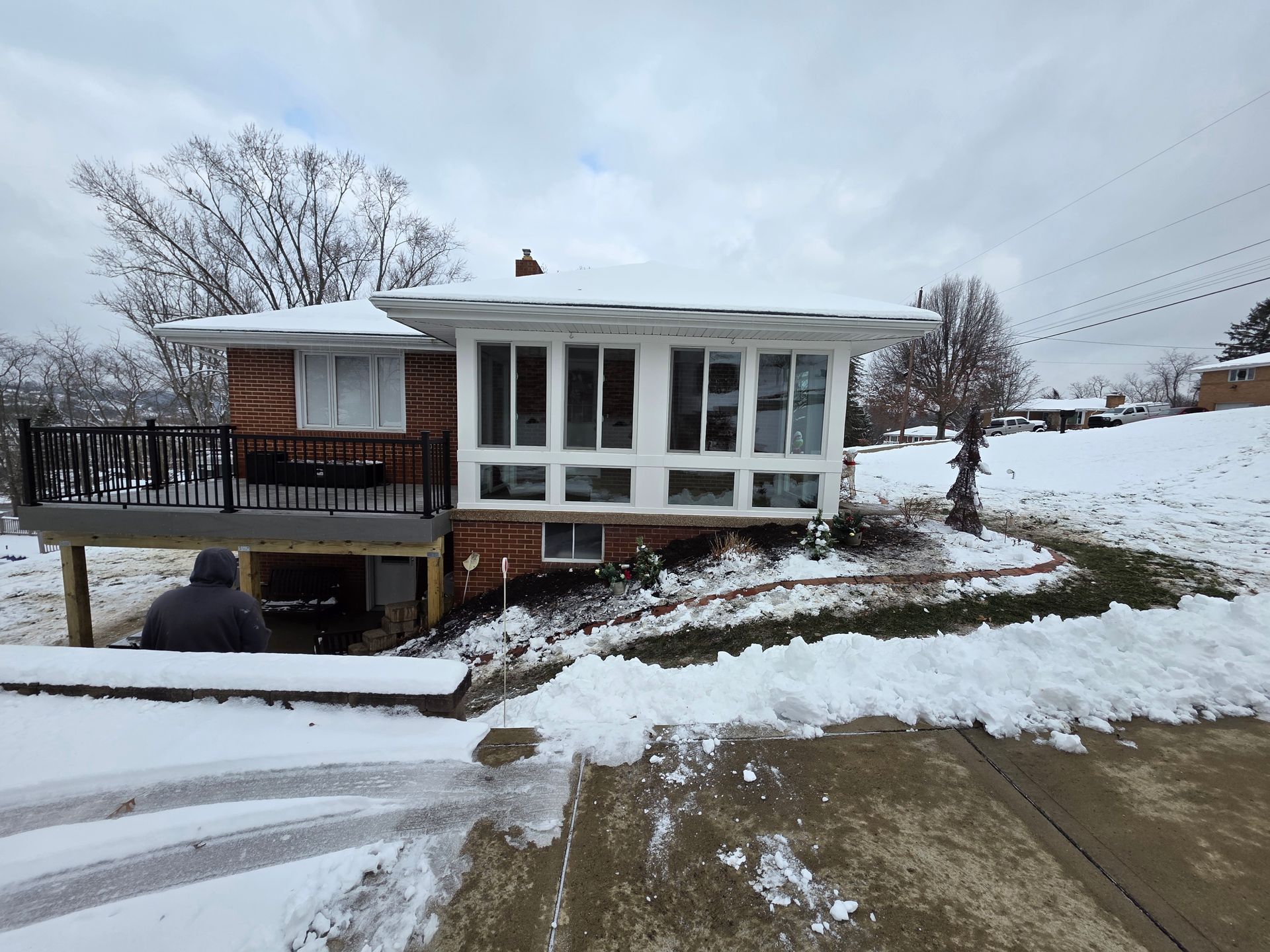 A house with a snow-covered yard and a deck. A person is sitting on the deck under the stairs.
