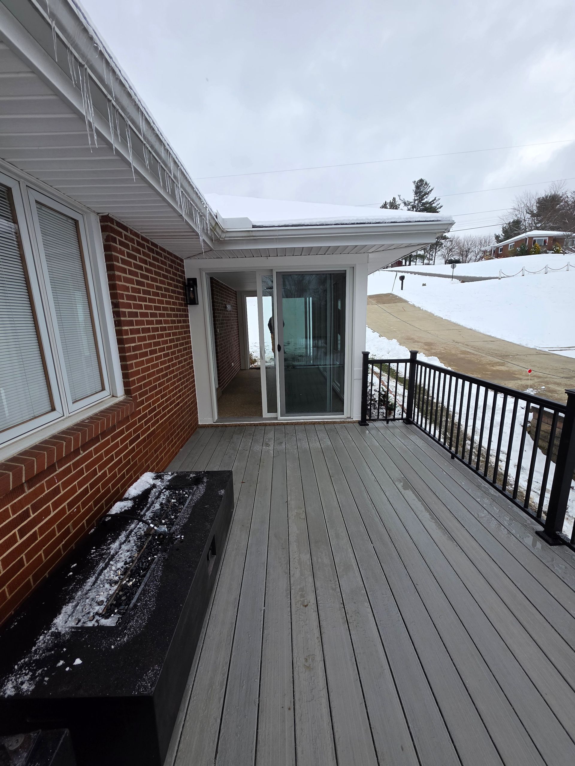 Snowy deck next to a brick house with sliding glass doors. Icicles hang from the roof.