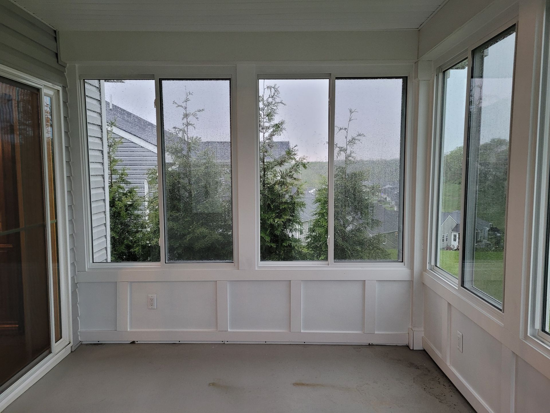 Sunroom with white paneling and large windows overlooking trees on a rainy day.