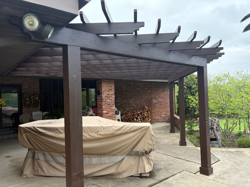 Brown pergola over a patio. A covered table sits in the foreground. Brick wall and trees are visible in the background.