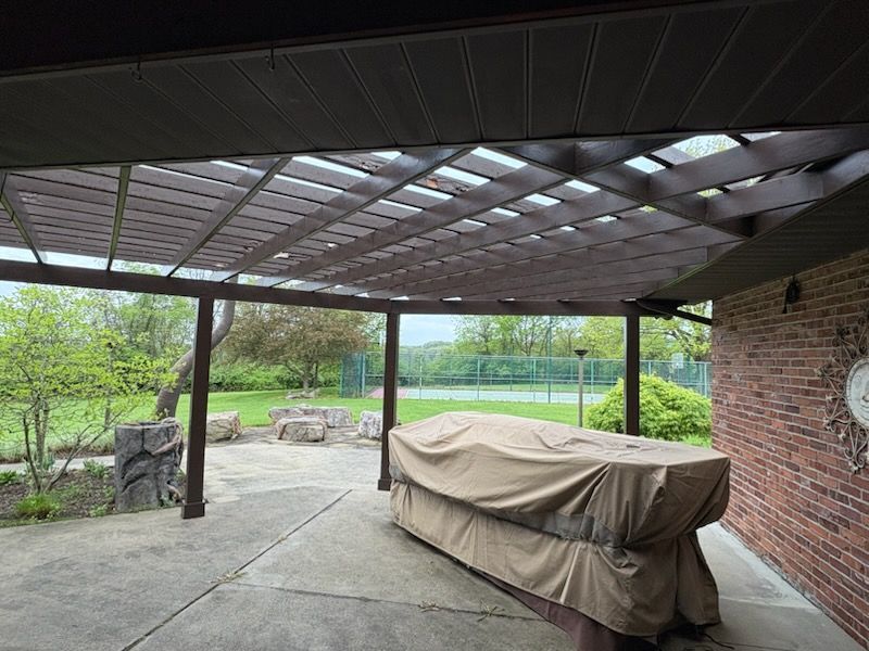 Patio with brown pergola, covered table, and view of green yard and tennis court.