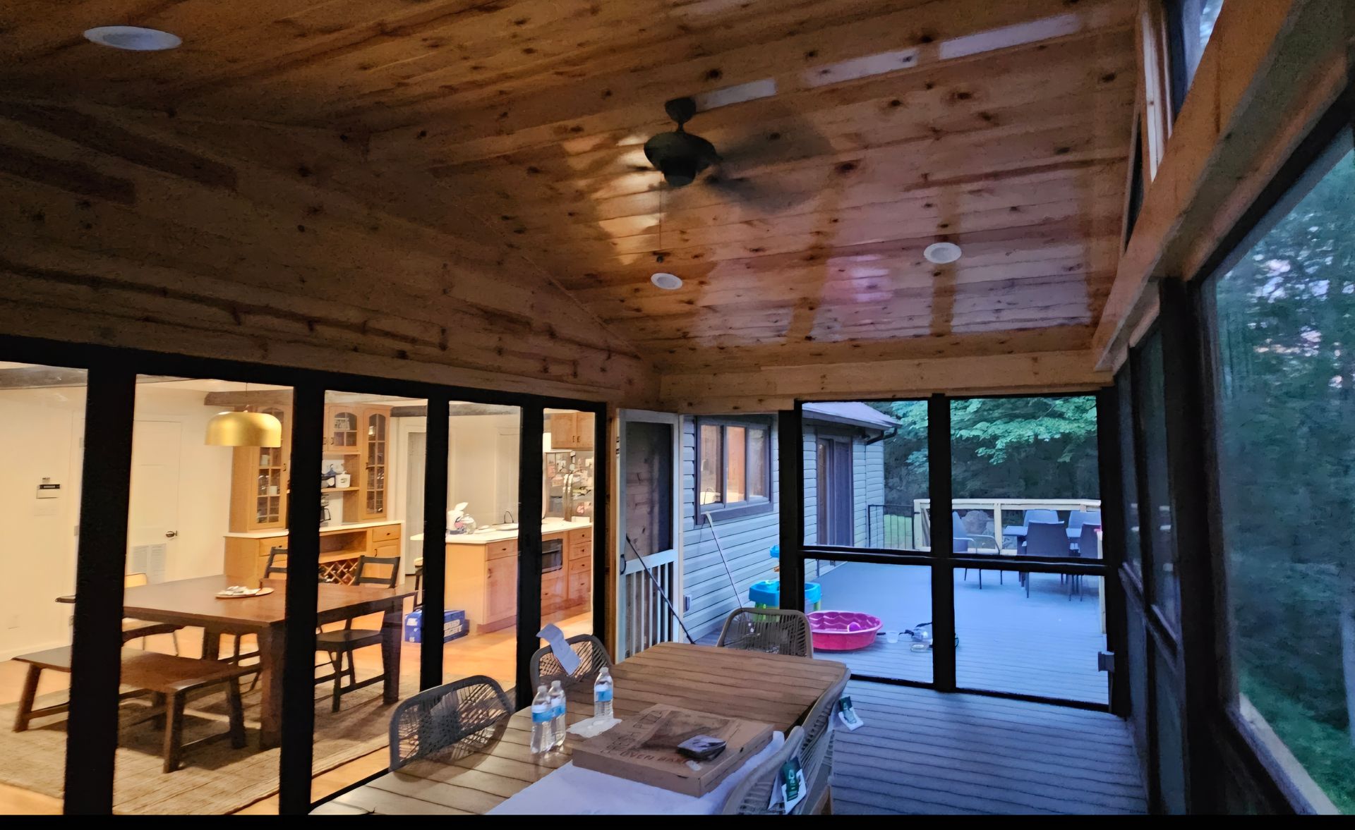 Enclosed porch with wood ceiling and glass walls, overlooking a deck and interior dining area.