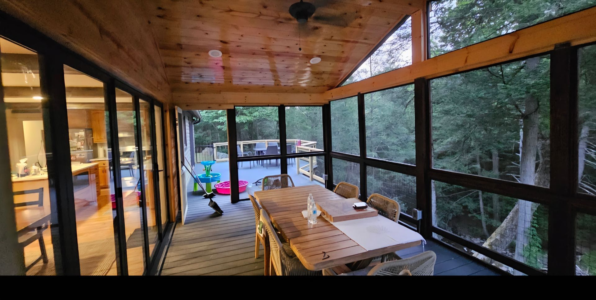 Screened-in porch with a wooden table and chairs, surrounded by windows overlooking a wooded area.