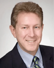 Man in suit smiling, light skin, short brown hair, blue shirt, green and blue striped tie.