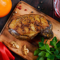 A seared foie gras steak on a wooden board with walnuts, fresh parsley, an orange, and red sauce in the background.