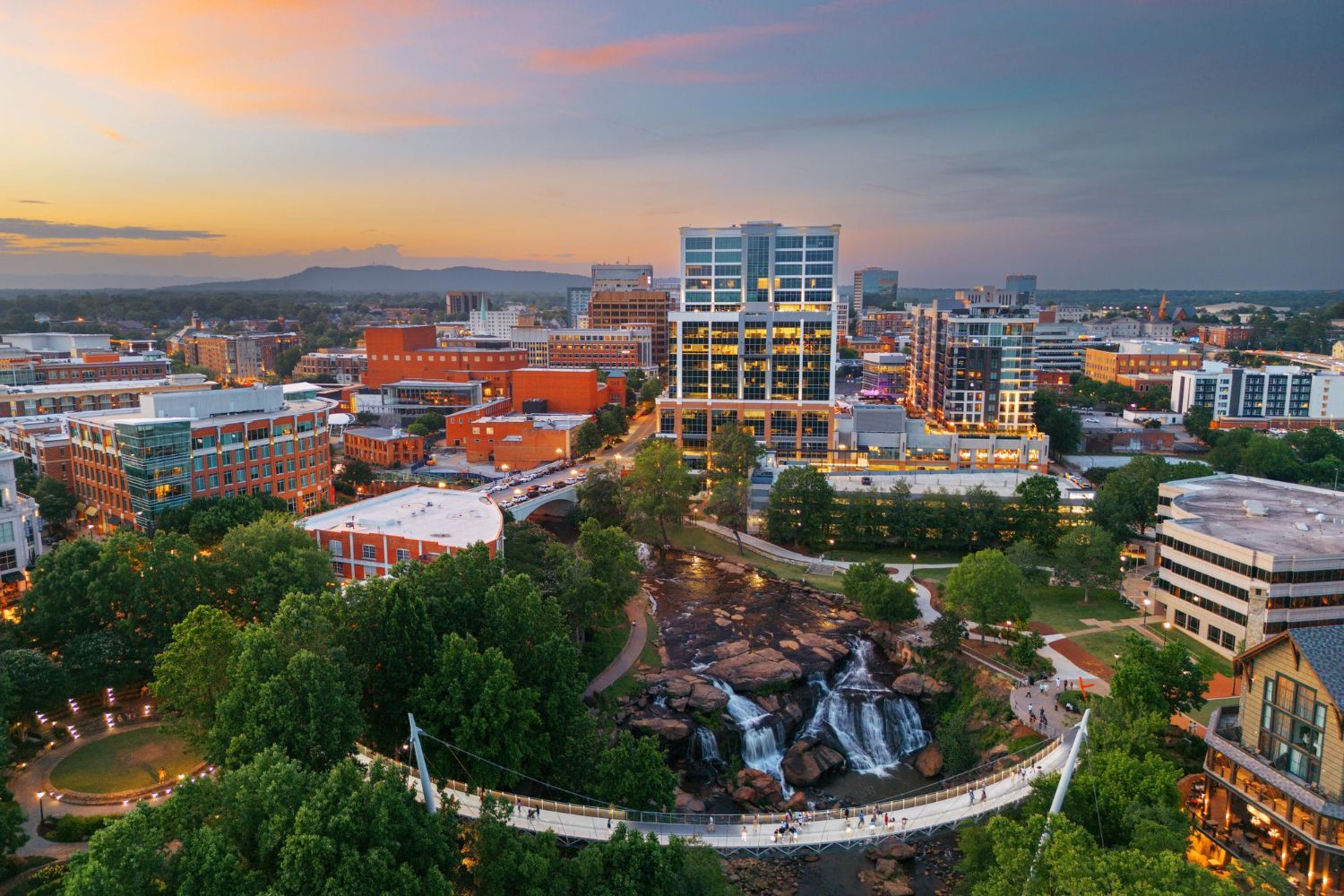 City skyline at sunset with trees, a park waterfall, and modern buildings against a colorful sky