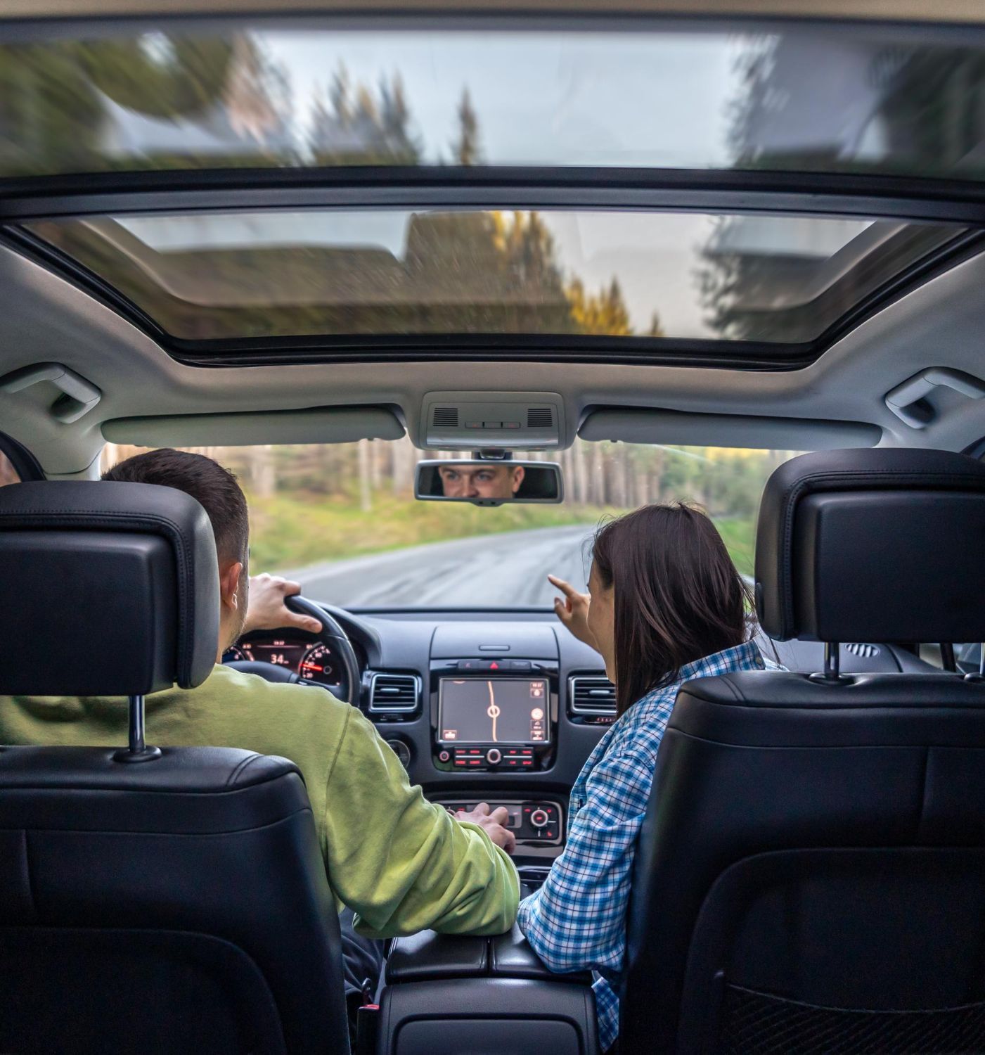 Two people sit in the front seat of a car, driving on a forest road with a panoramic sunroof.
