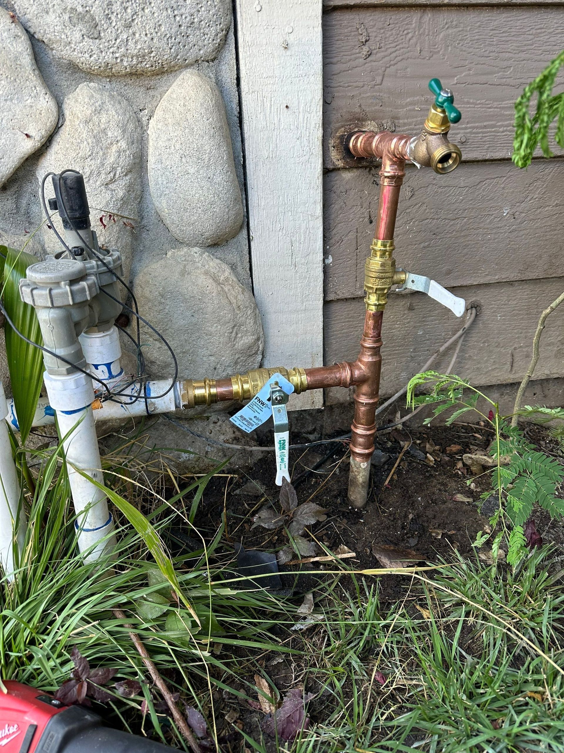 Outdoor water pipes with faucets and valves beside a stone wall and garden bed.