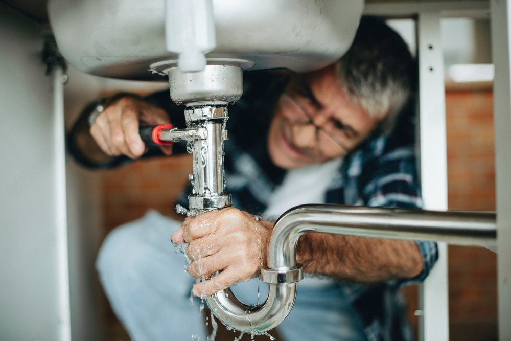 Plumber tightening pipes under a sink with a wrench, repairing a kitchen drain.