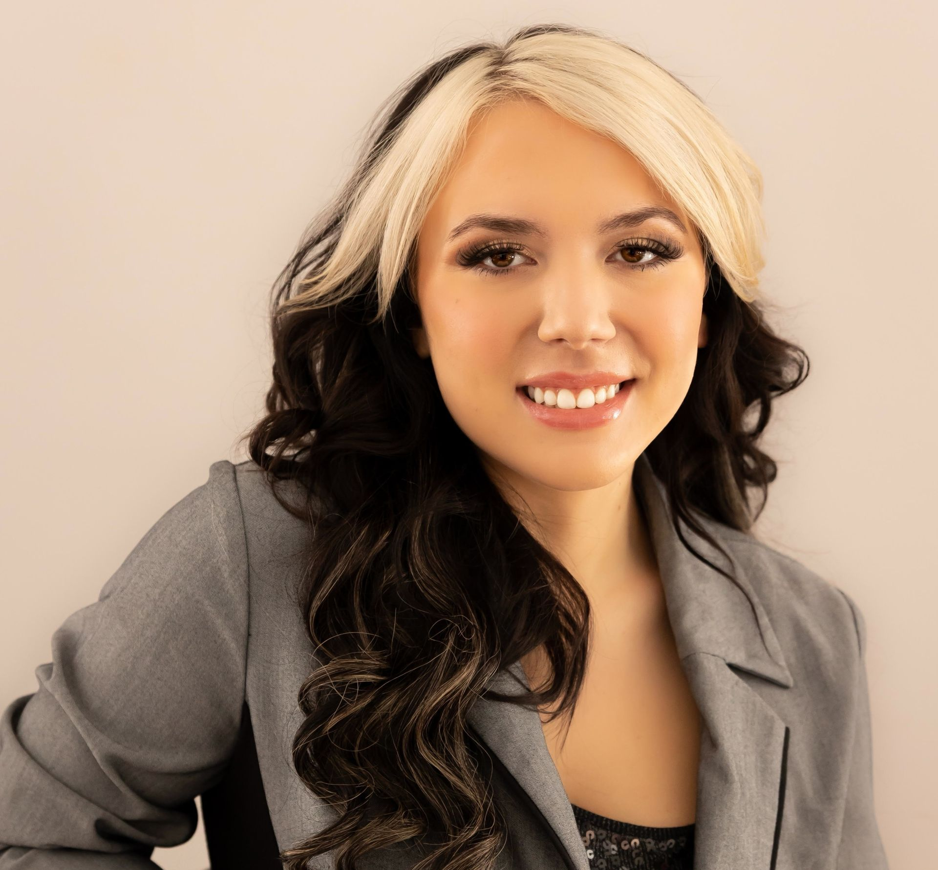 Smiling woman with two-toned dark and blonde hair wearing a grey blazer against a neutral backdrop.