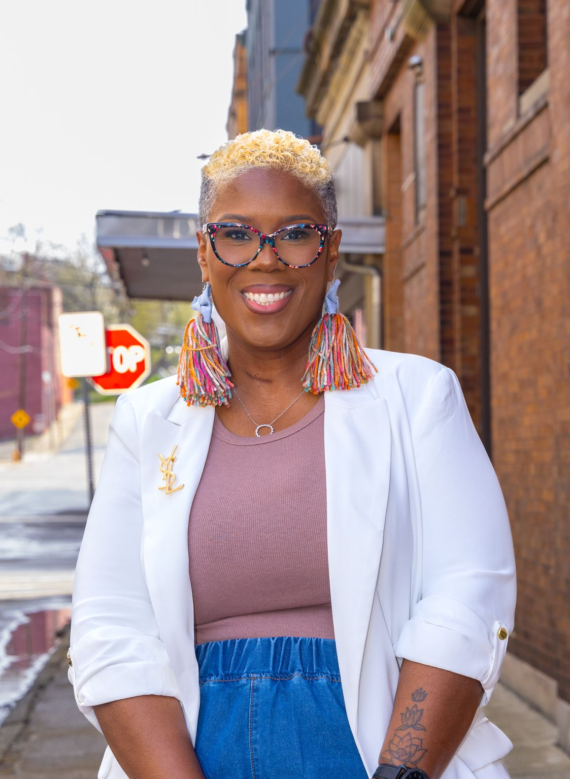 A person with short blonde hair and glasses wearing a white blazer, mauve top, and denim skirt stands on a city street.