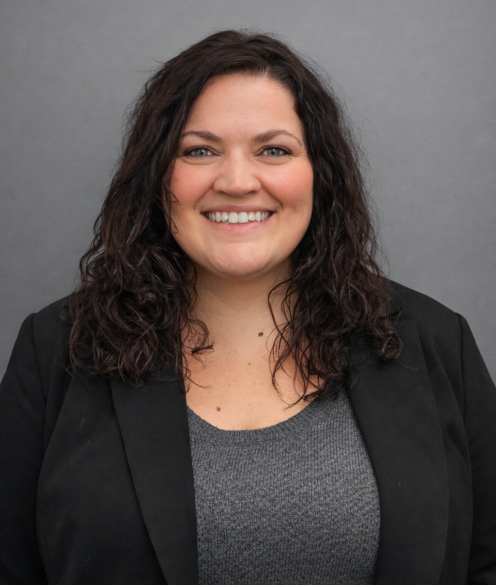 Smiling person with dark curly hair wearing a gray top and black blazer against a gray background.