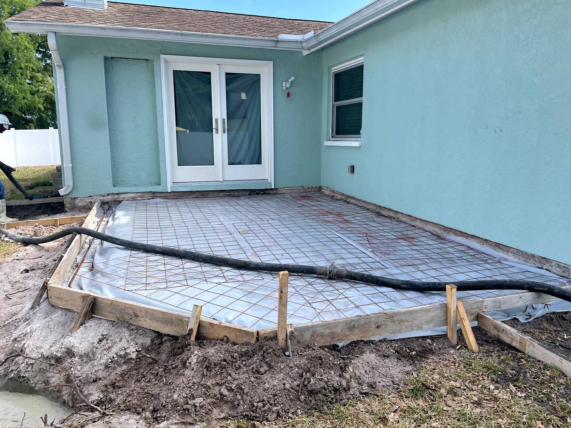 Concrete patio construction site, wood frame, rebar, plastic covering, next to a teal house with glass doors.