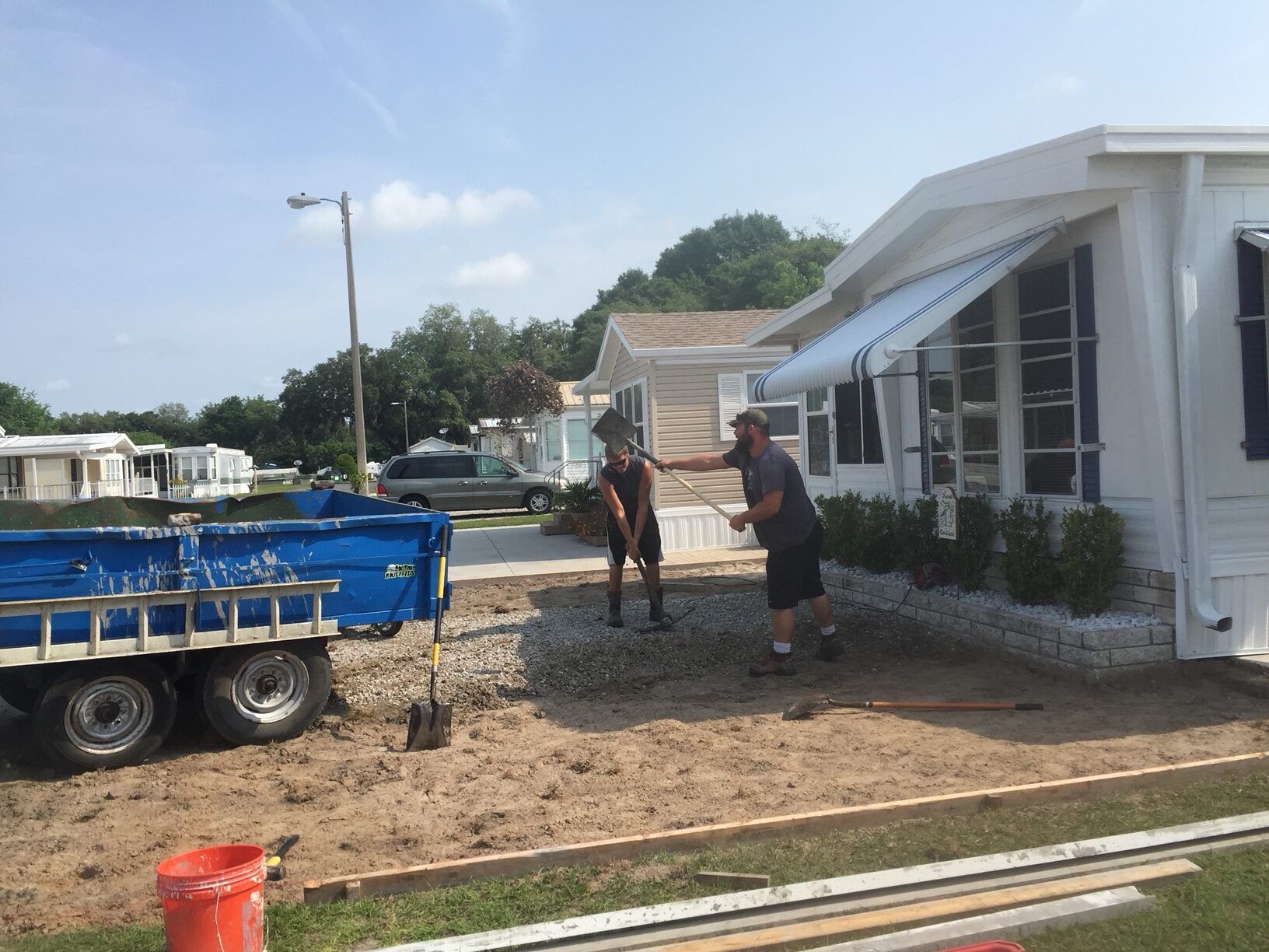 Two men working on landscaping next to a white mobile home with blue trailer and sunny sky.