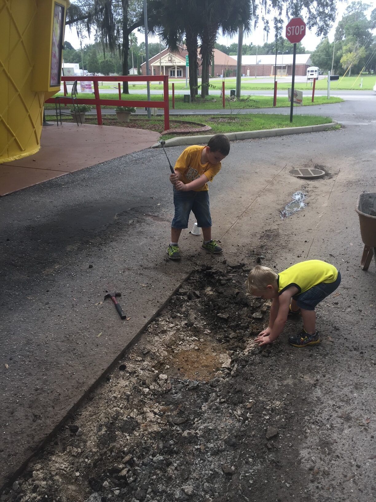 Two young boys in yellow shirts digging in asphalt. One swings a hammer, other kneels.