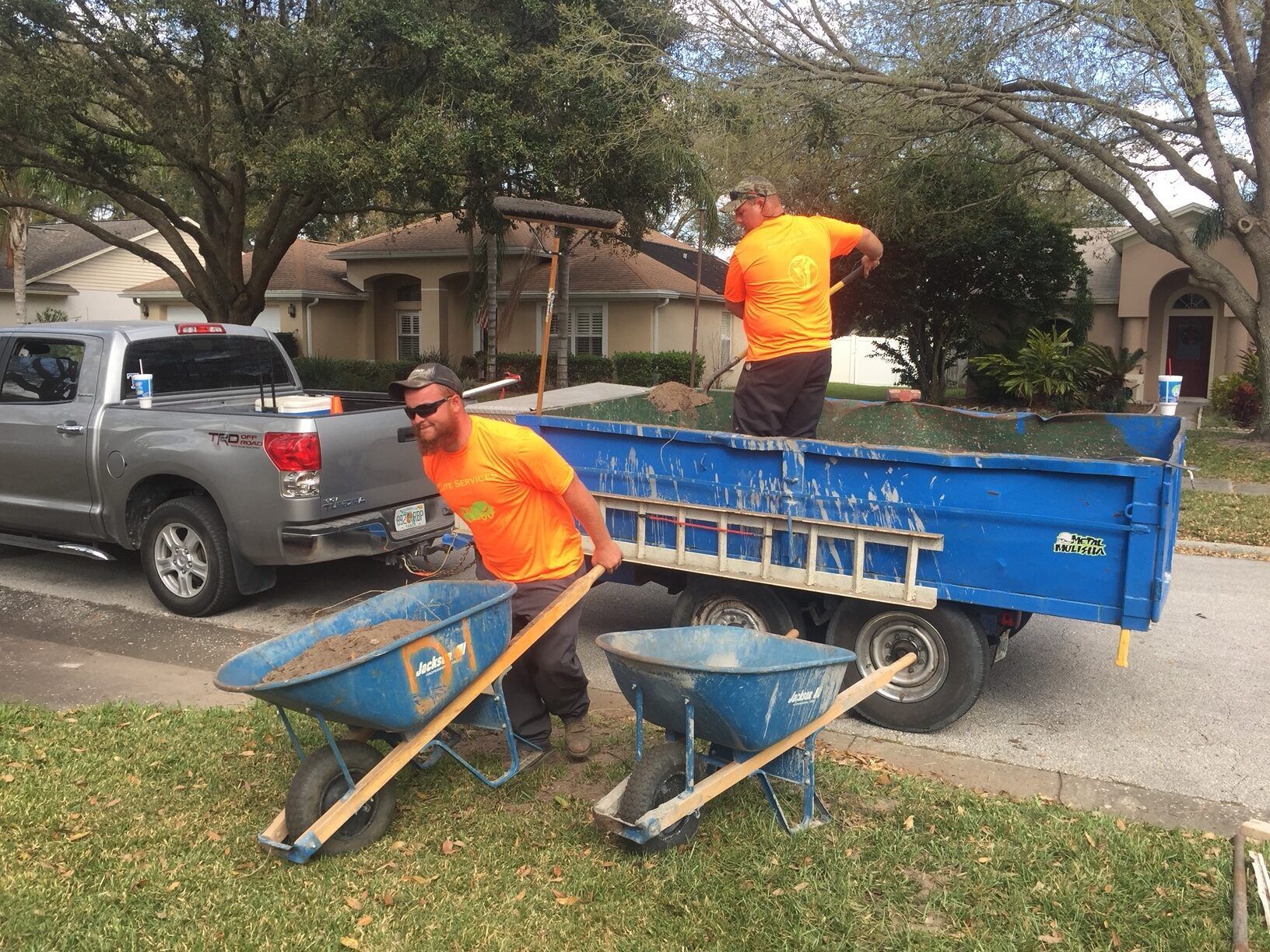 Two workers in orange shirts loading soil into a trailer. One pushes a wheelbarrow, the other shovels.