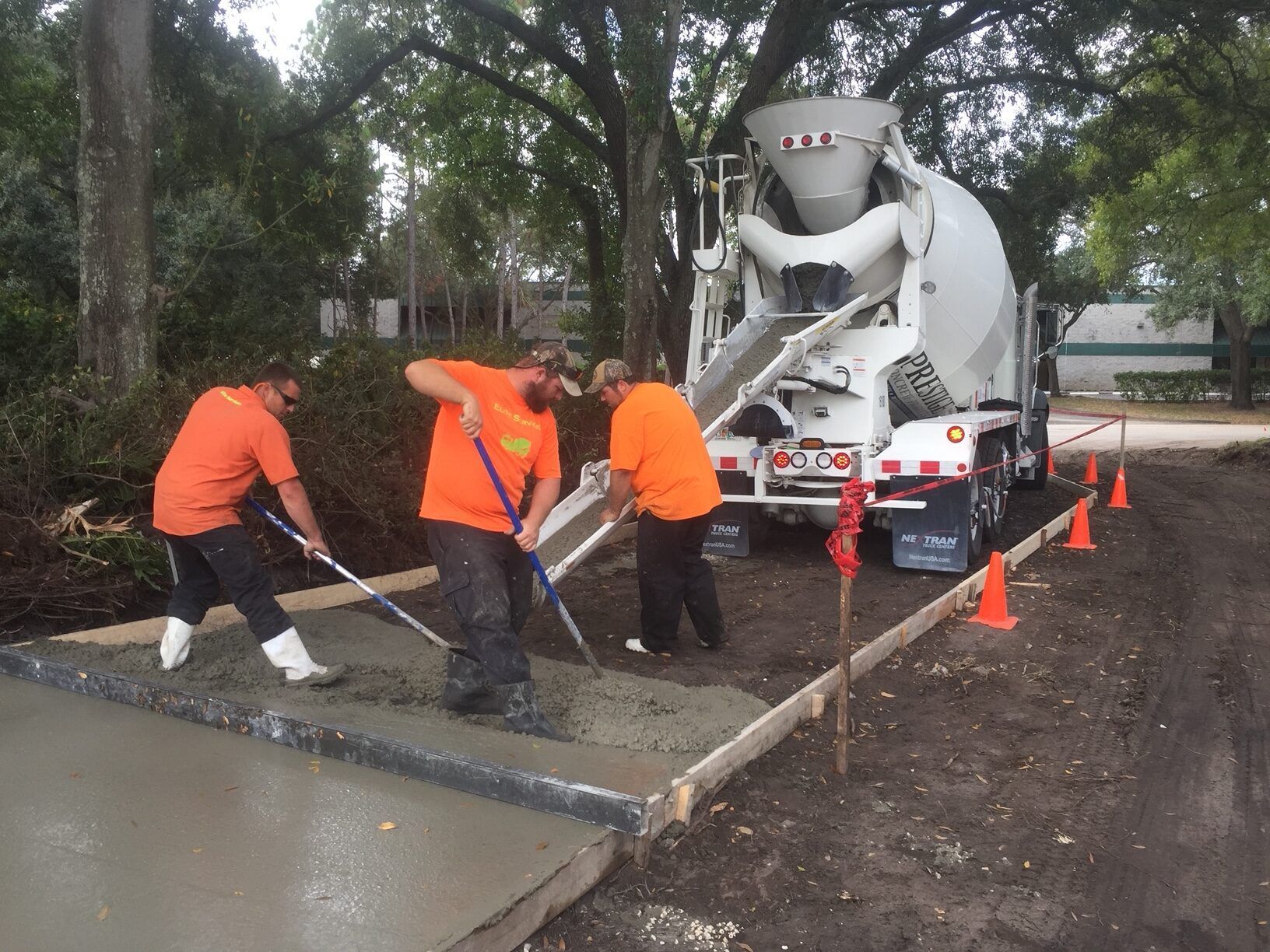 Construction workers in orange shirts pouring concrete from a truck into a framed area; outdoors.