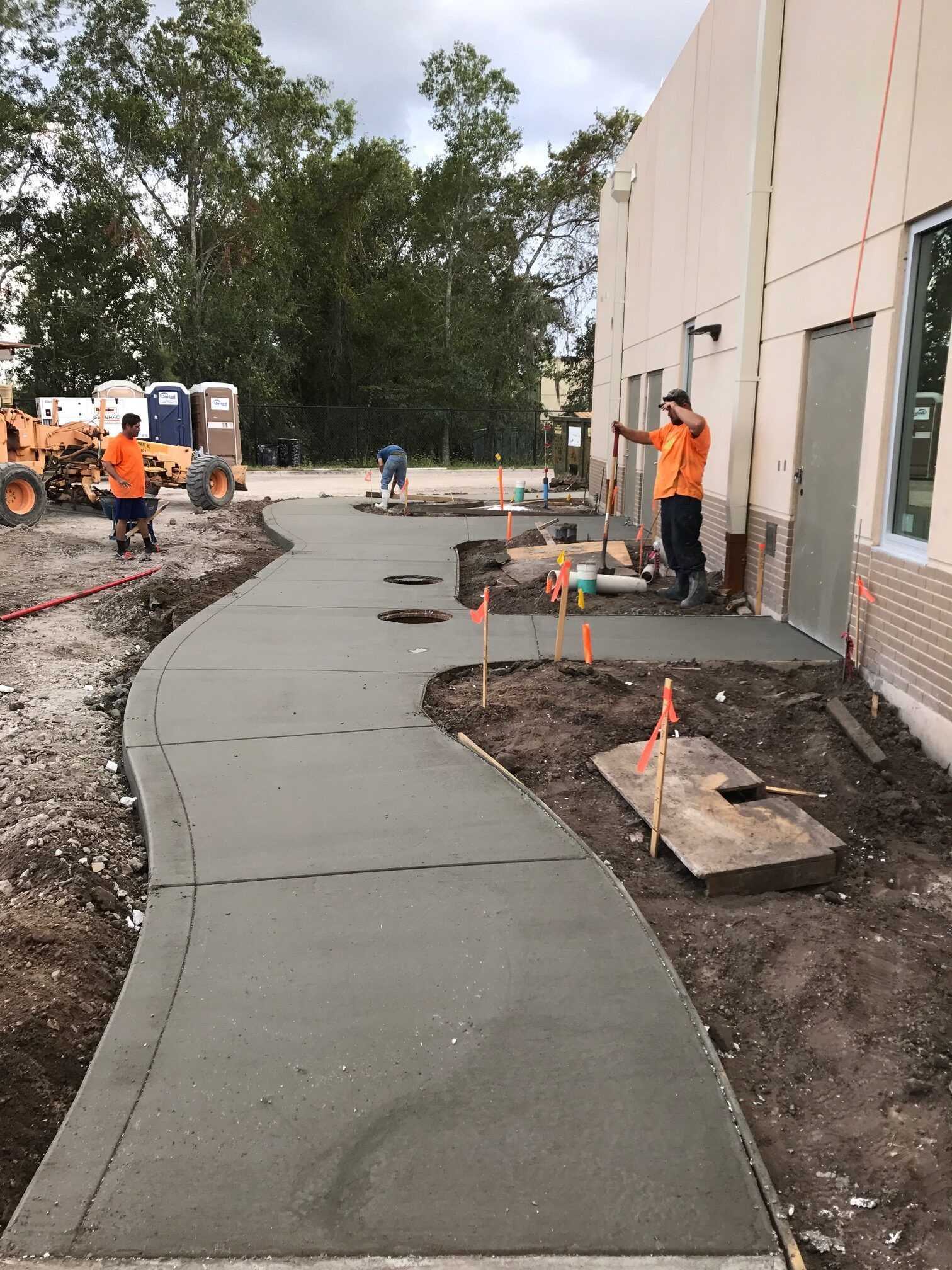 Workers pouring concrete sidewalk outside a building. Two workers in orange vests, cloudy sky.