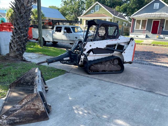 White Bobcat skid steer on a sidewalk with a pickup truck parked behind it, houses in background.