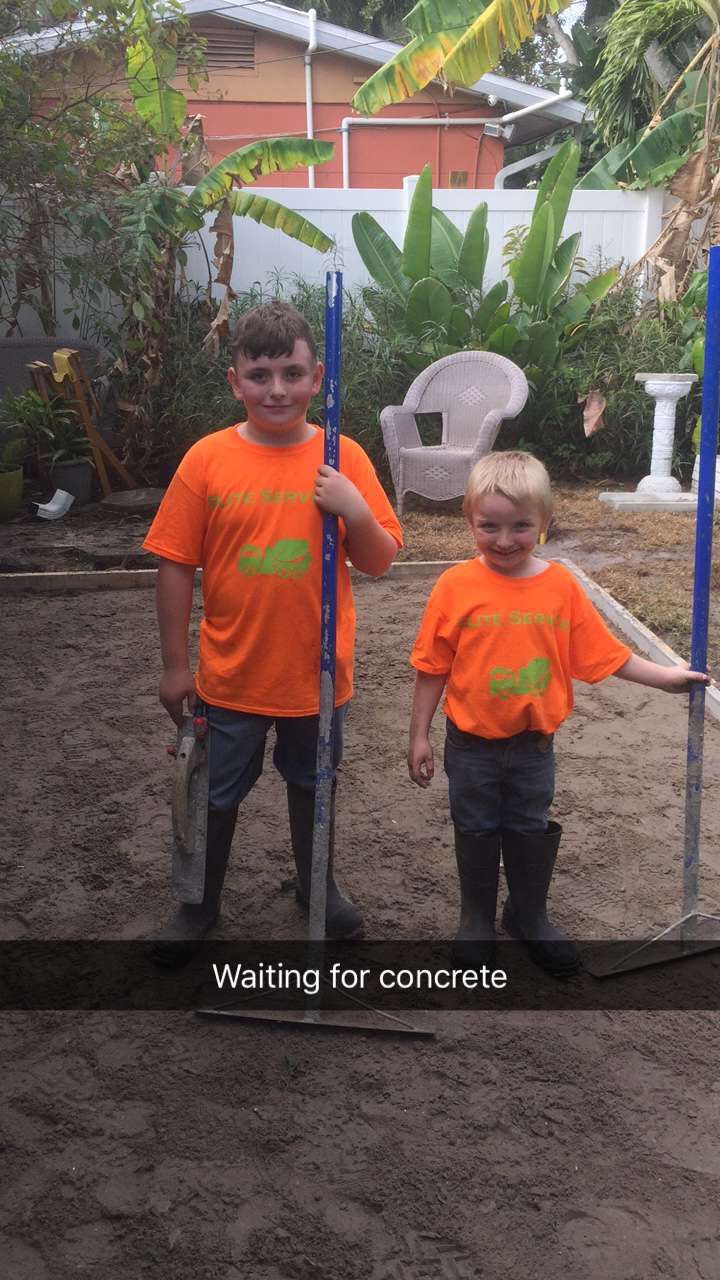 Two boys in orange shirts stand in dirt, holding poles, waiting for concrete.
