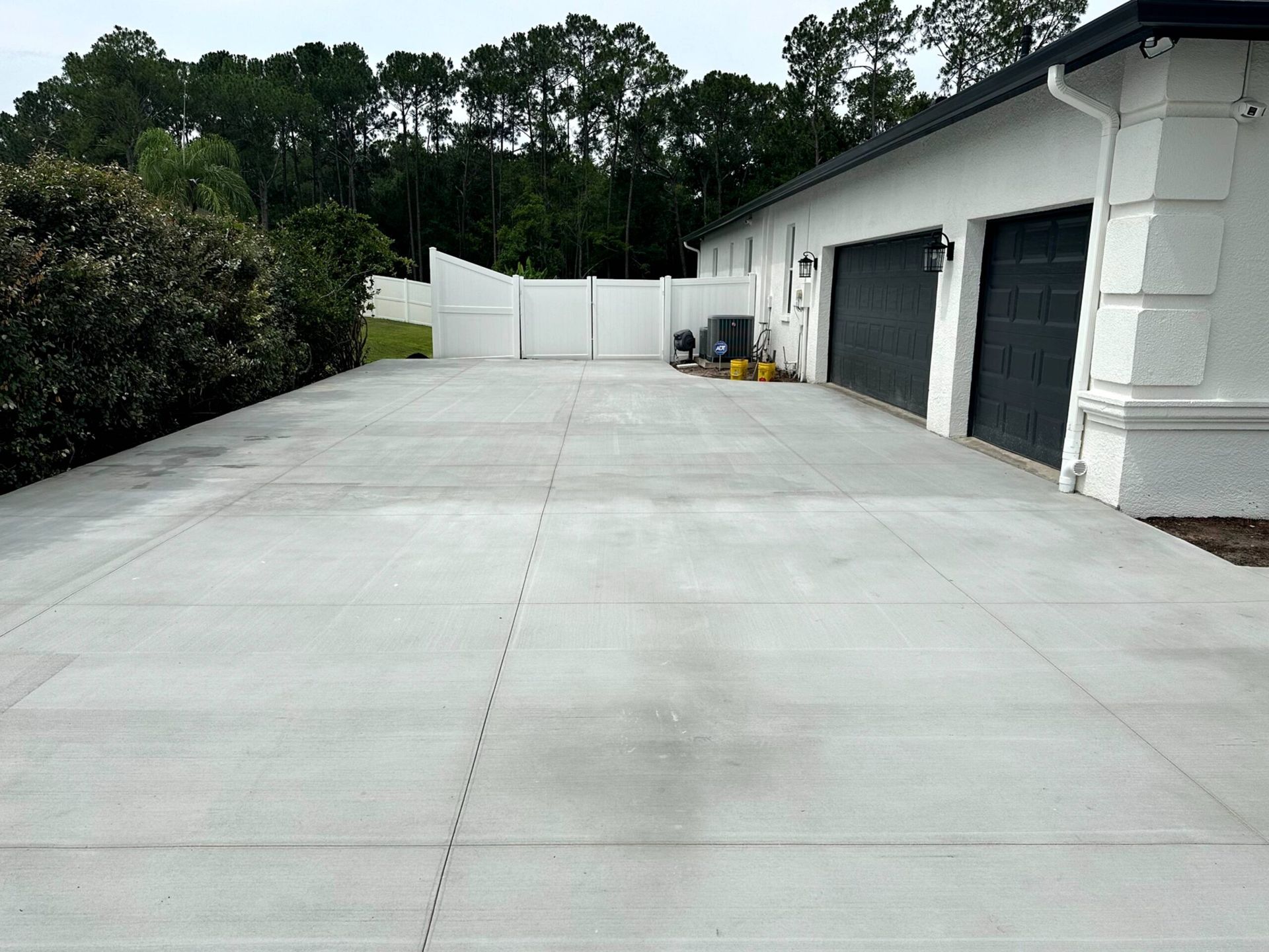 Gray concrete driveway leading to white house with black garage doors and a white fence.