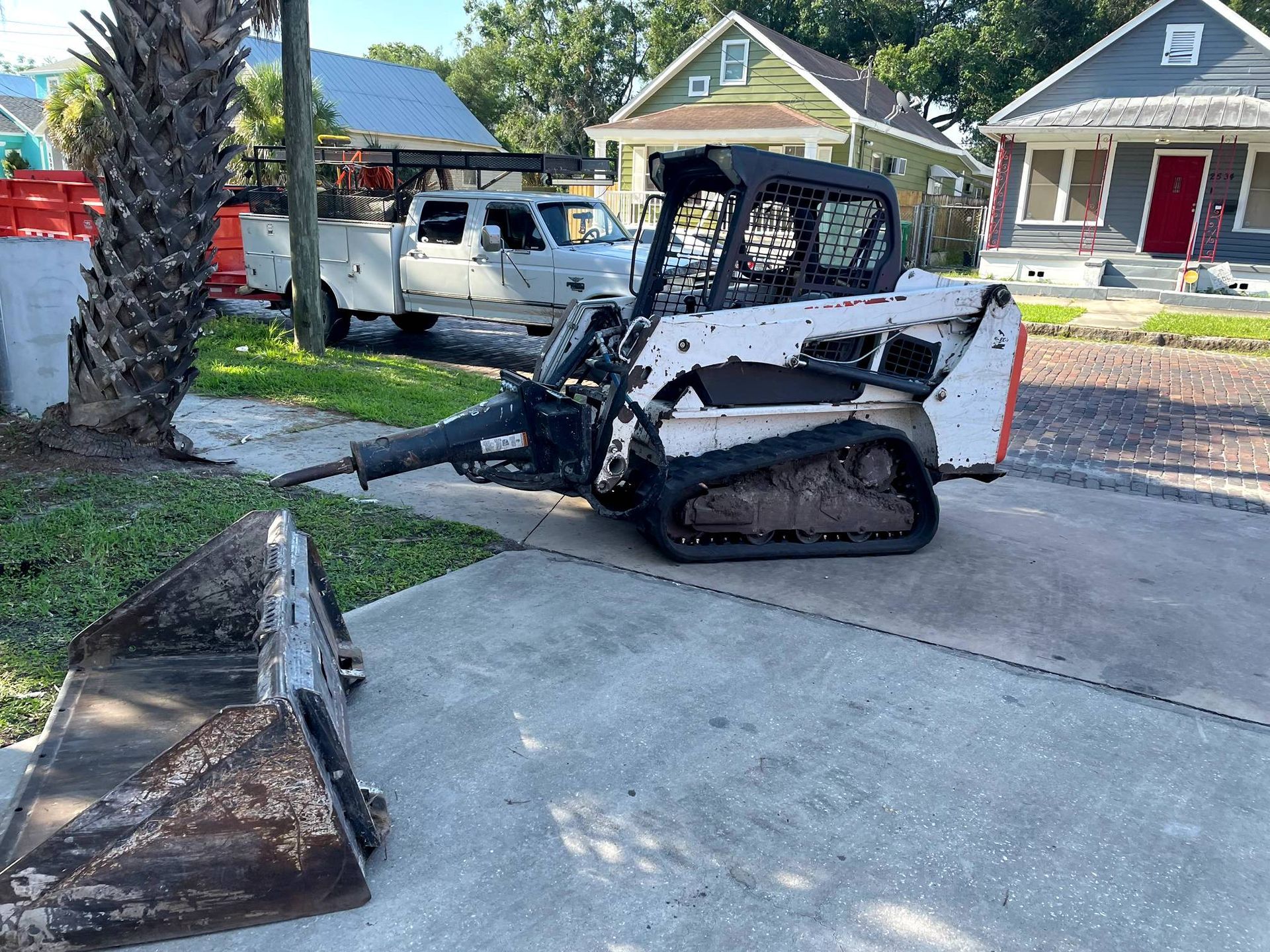 White Bobcat track loader with jackhammer attachment on a paved driveway next to a pickup truck and houses.