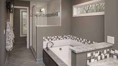 A modern bathroom featuring a white soaking tub with a tiled surround, grey walls, a tiled floor, and a walk-in shower.