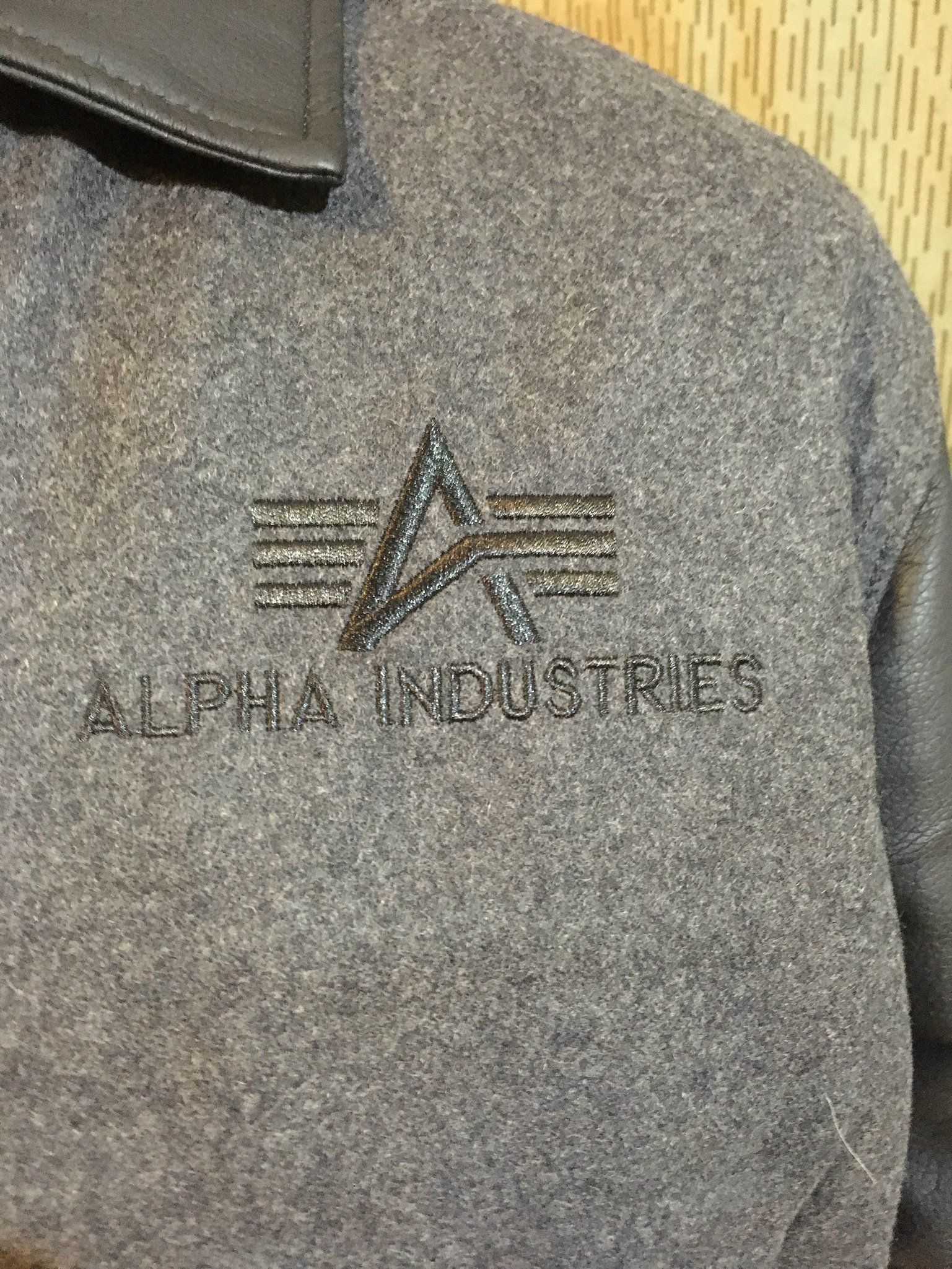 Graue Alpha Industries Jacke mit gesticktem Logo und Lederkragen.