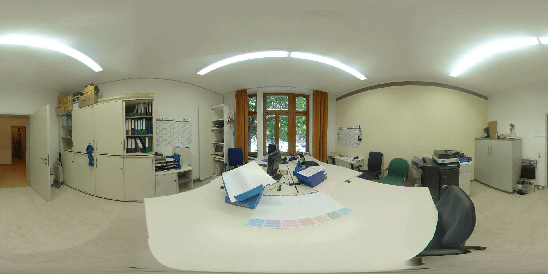 Ein Bürointerieur mit einem großen Schreibtisch, Aktenschränken und einem Fenster, durch das man Grünpflanzen sehen kann.