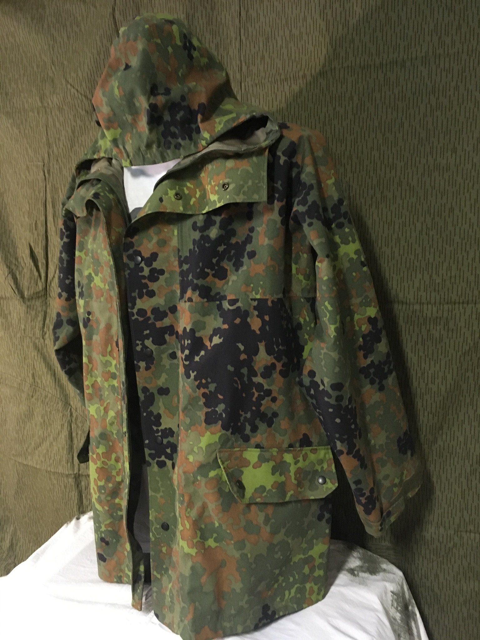 Eine Tarnjacke und Kapuze mit grünen, braunen und schwarzen Flecken vor einem ähnlichen Hintergrund.