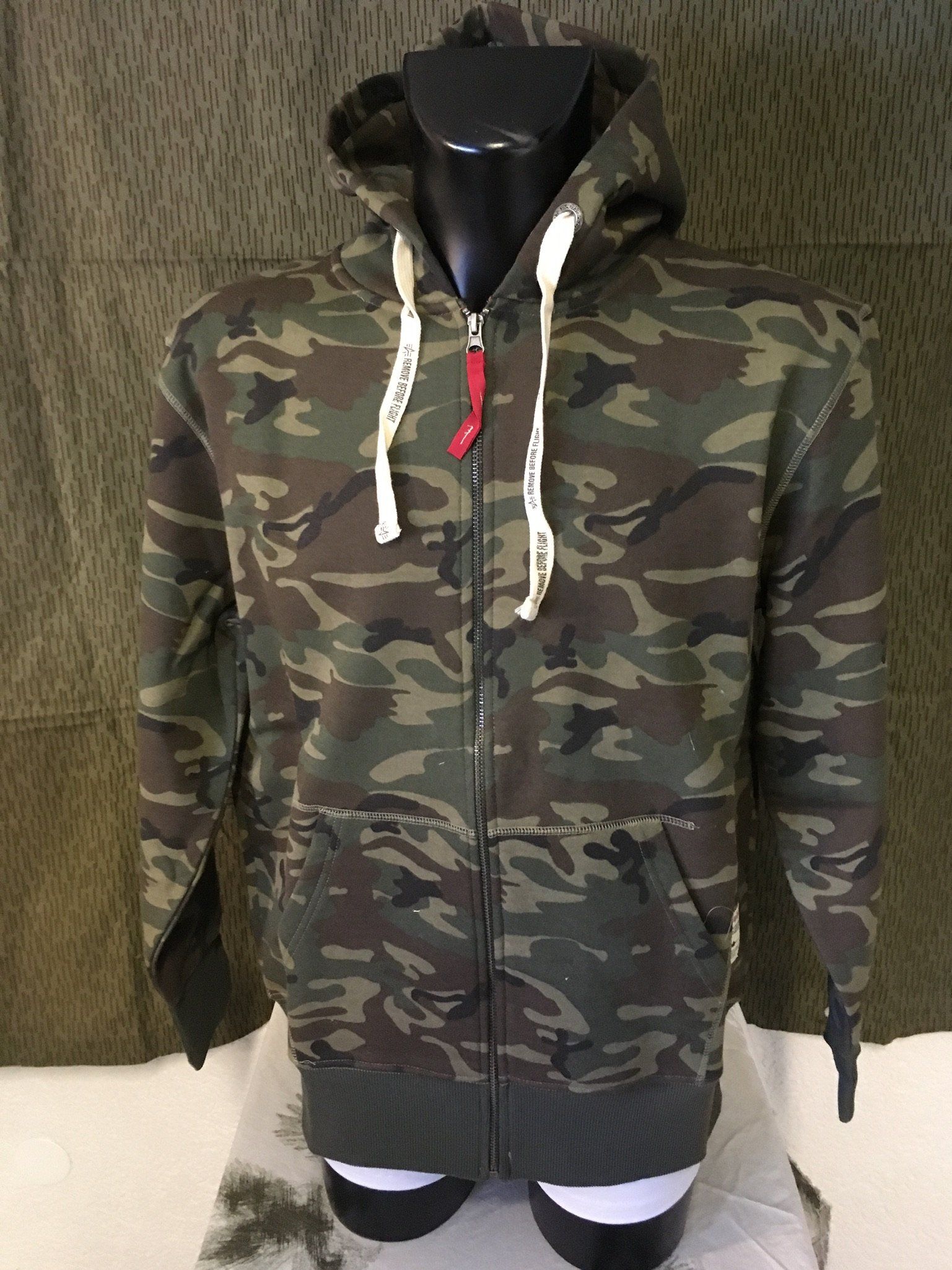 Camouflage-Kapuzenpullover mit Reißverschluss an einer Schaufensterpuppe, mit weißen Kordeln und einem roten Akzent.
