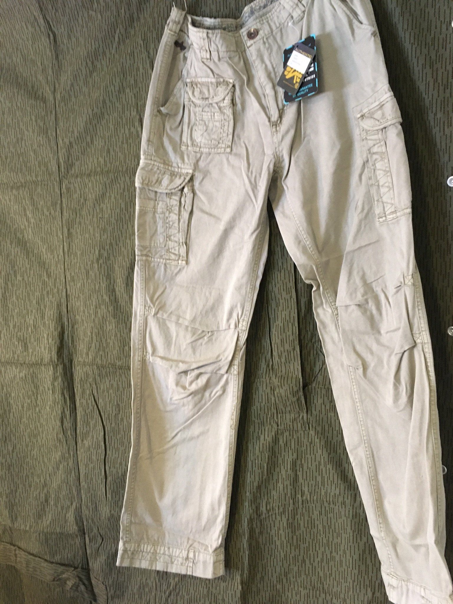 Beige Cargohose mit mehreren Taschen, noch mit Preisschild, auf grünem Hintergrund.