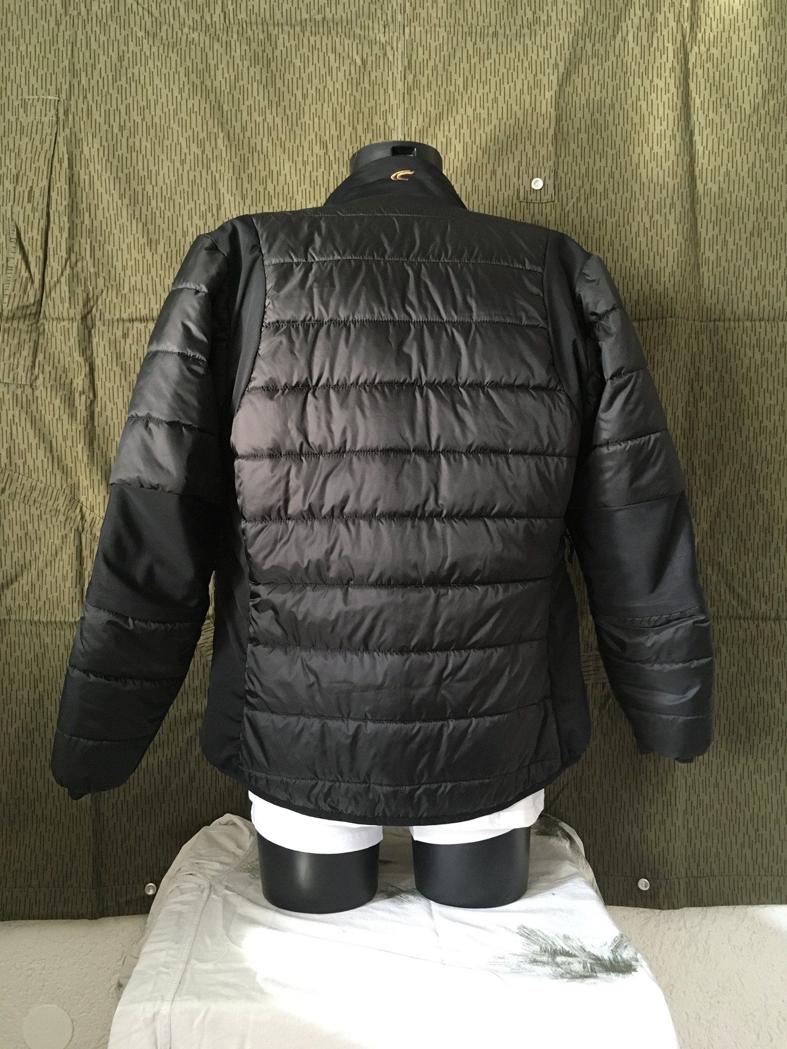 Schwarze Steppjacke auf einer Schaufensterpuppe, mit grünem Camouflage-Hintergrund.