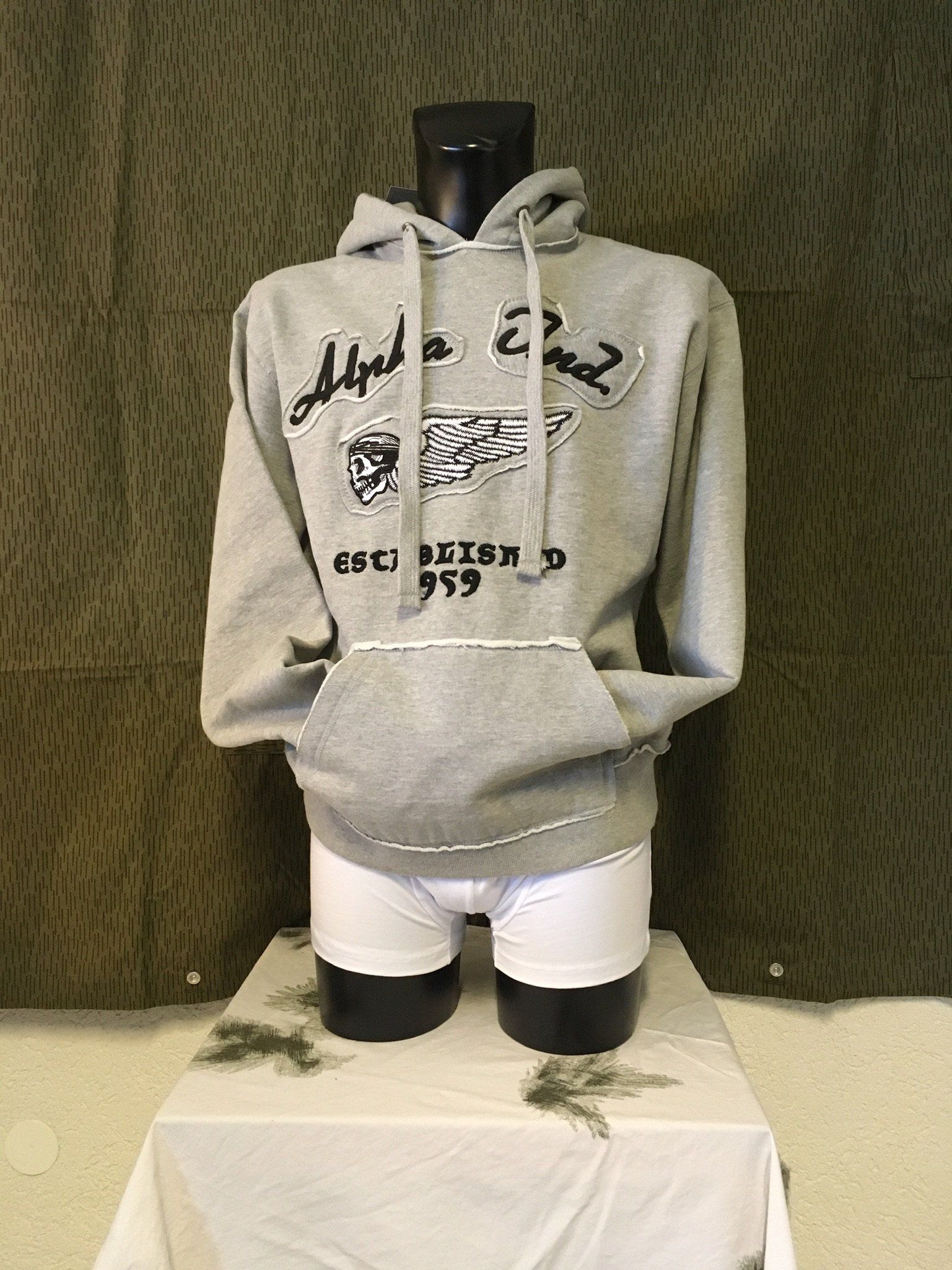 Grauer Kapuzenpullover an einer Schaufensterpuppe mit dem Text „Alpha Phi Delta“ vor einem Tarnhintergrund.