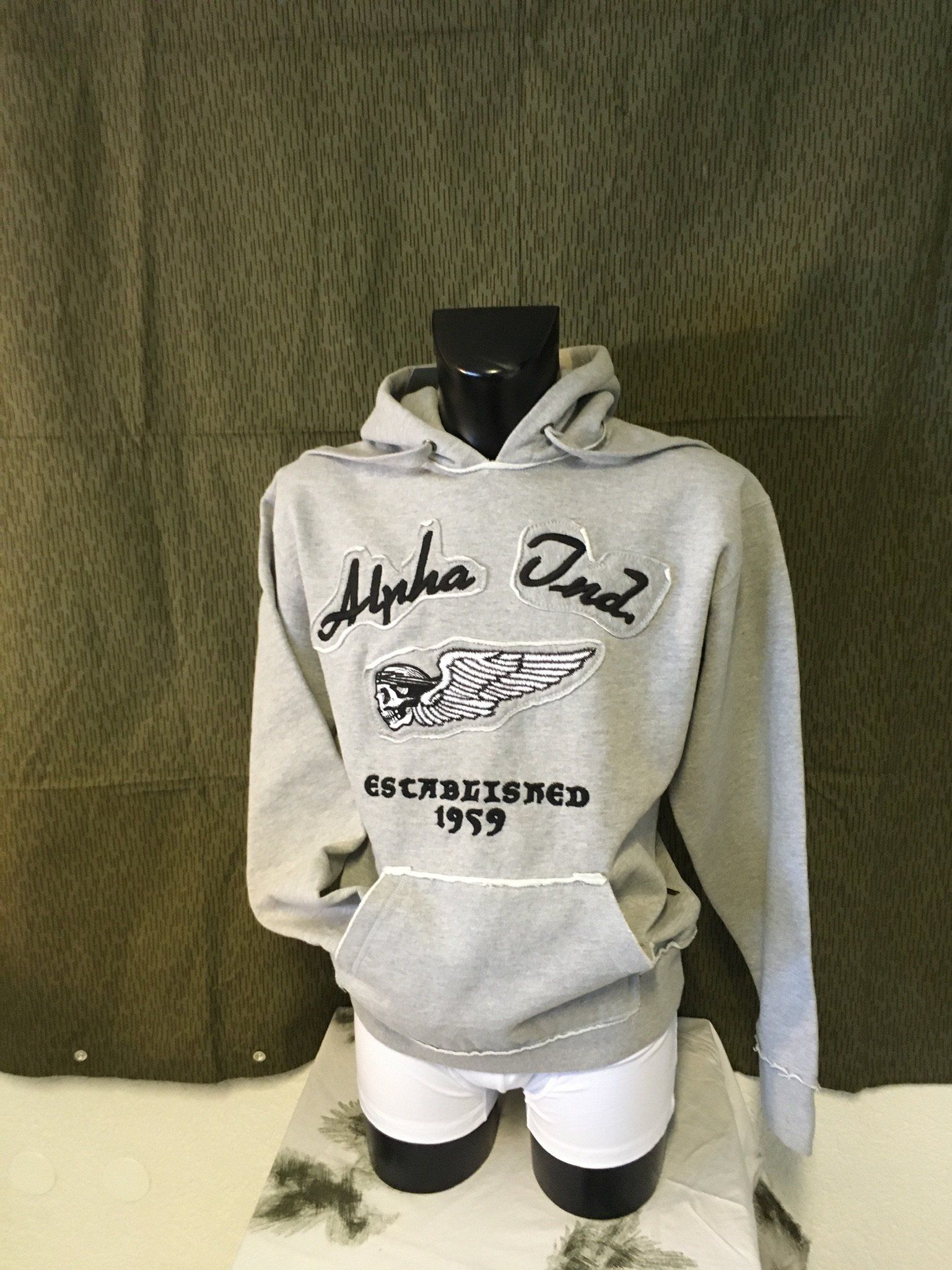Grauer Kapuzenpullover an einer Schaufensterpuppe mit „Alpha One“-Logo und Tasche. Gegründet 1969.