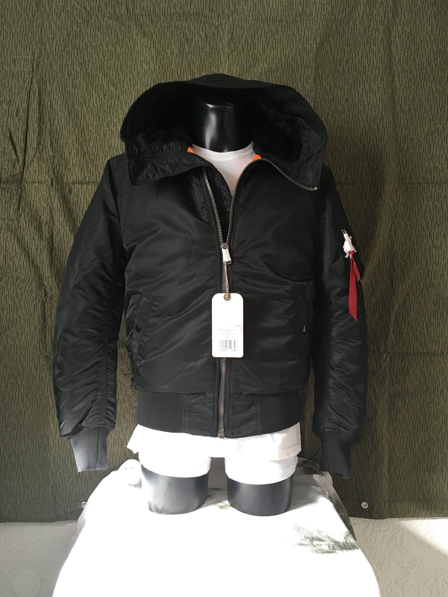 Schwarze Bomberjacke mit Kunstpelzkapuze, auf einer Schaufensterpuppe, vor einem Tarnhintergrund.