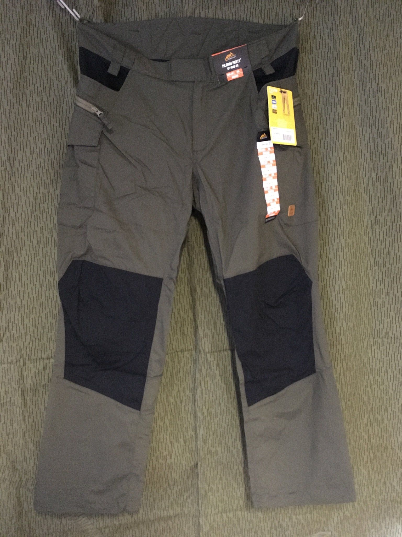 Graue Arbeitshose mit schwarzen Akzenten an Knien und Taschen, hängt auf einem Kleiderbügel mit Etiketten.