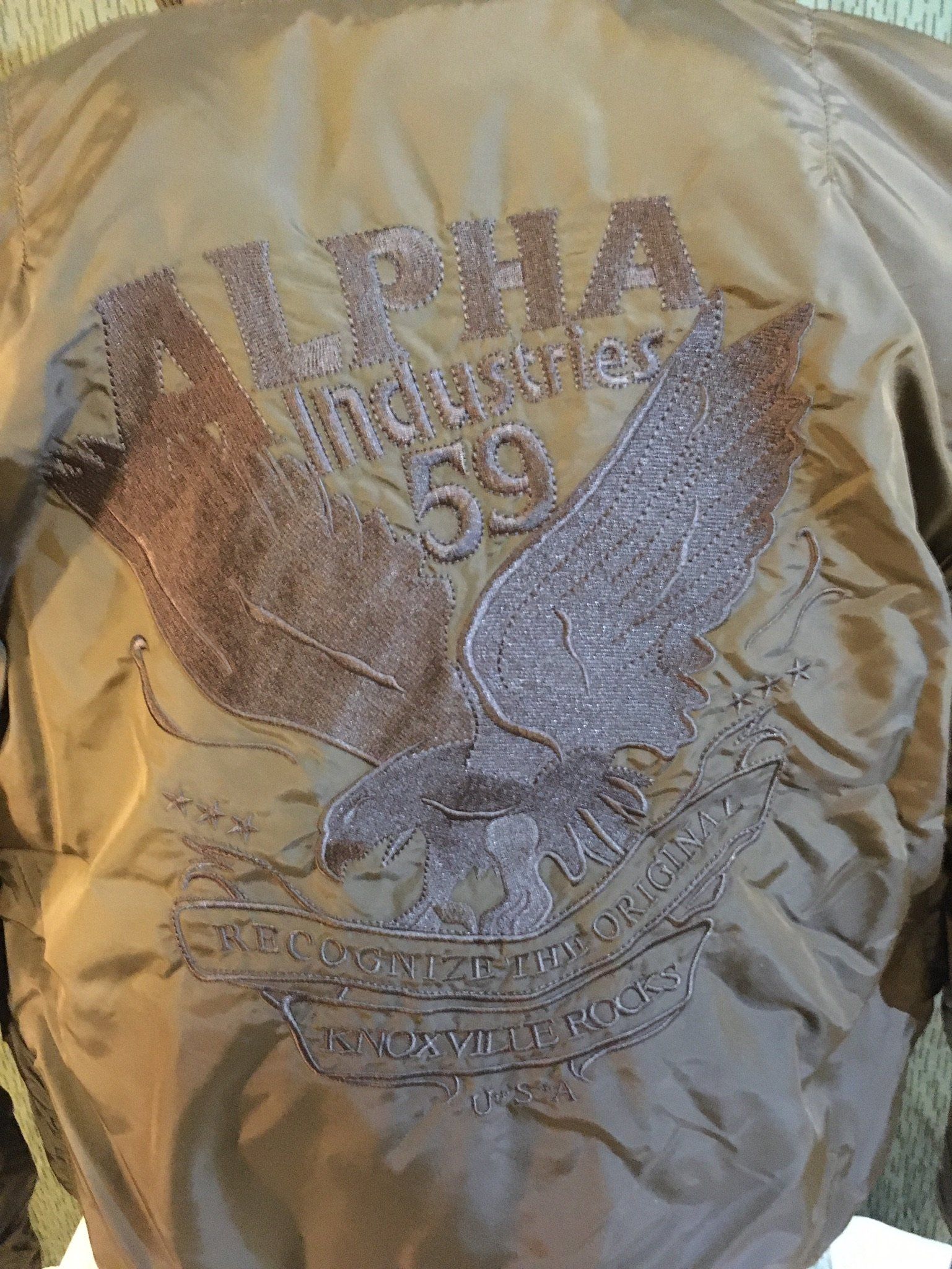Rückseite einer olivgrünen Alpha Industries Jacke mit gesticktem Logo und Adlergrafik.