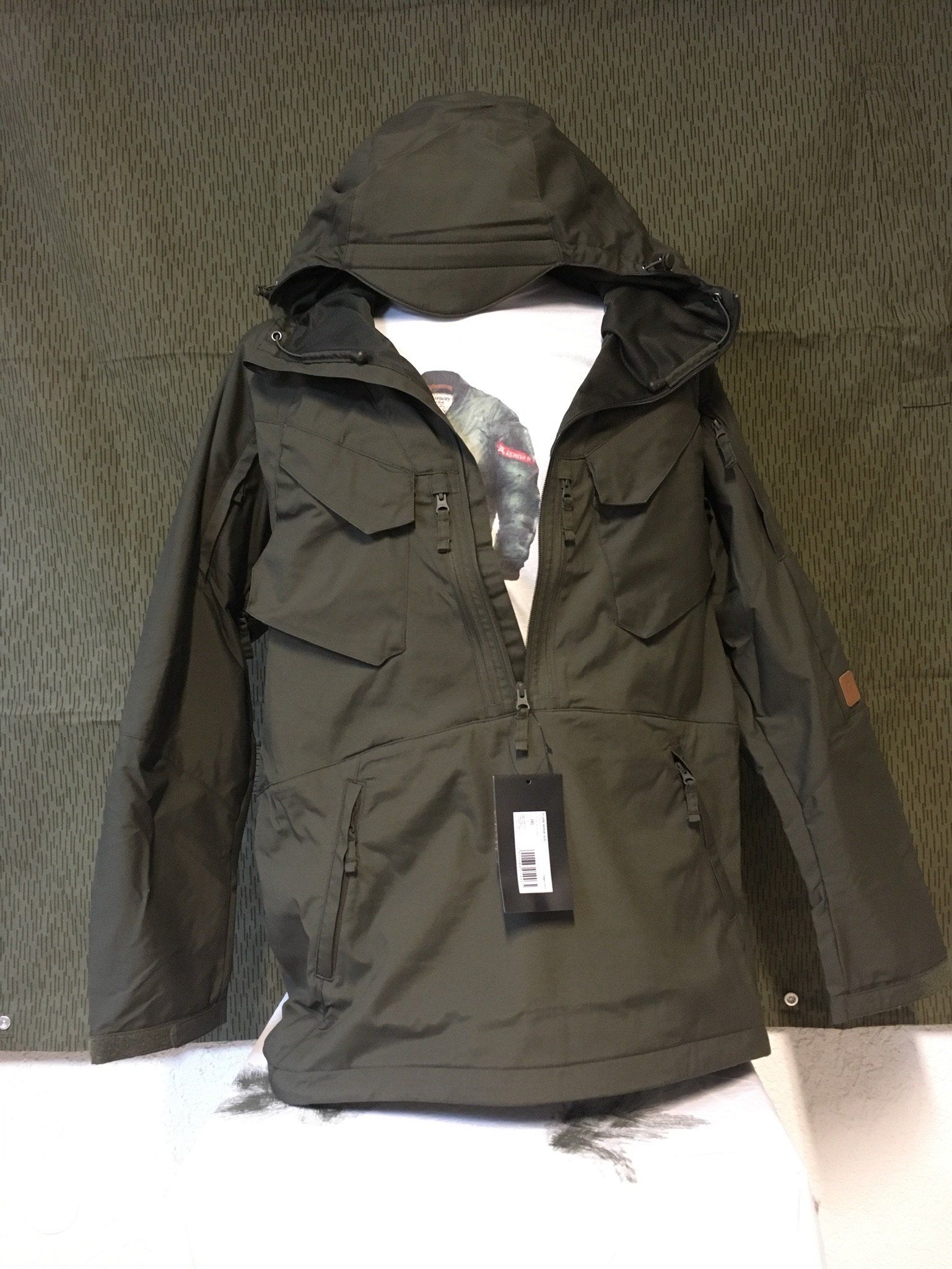 Grüne Jacke im Anorak-Stil mit Kapuze, getragen über einem weißen Hemd, auf strukturiertem Hintergrund.