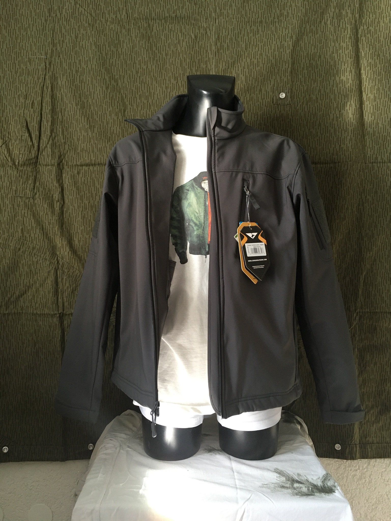 Eine dunkelgraue Jacke an einer Schaufensterpuppe, über einem weißen T-Shirt, vor einem grün-weißen Tarnhintergrund.