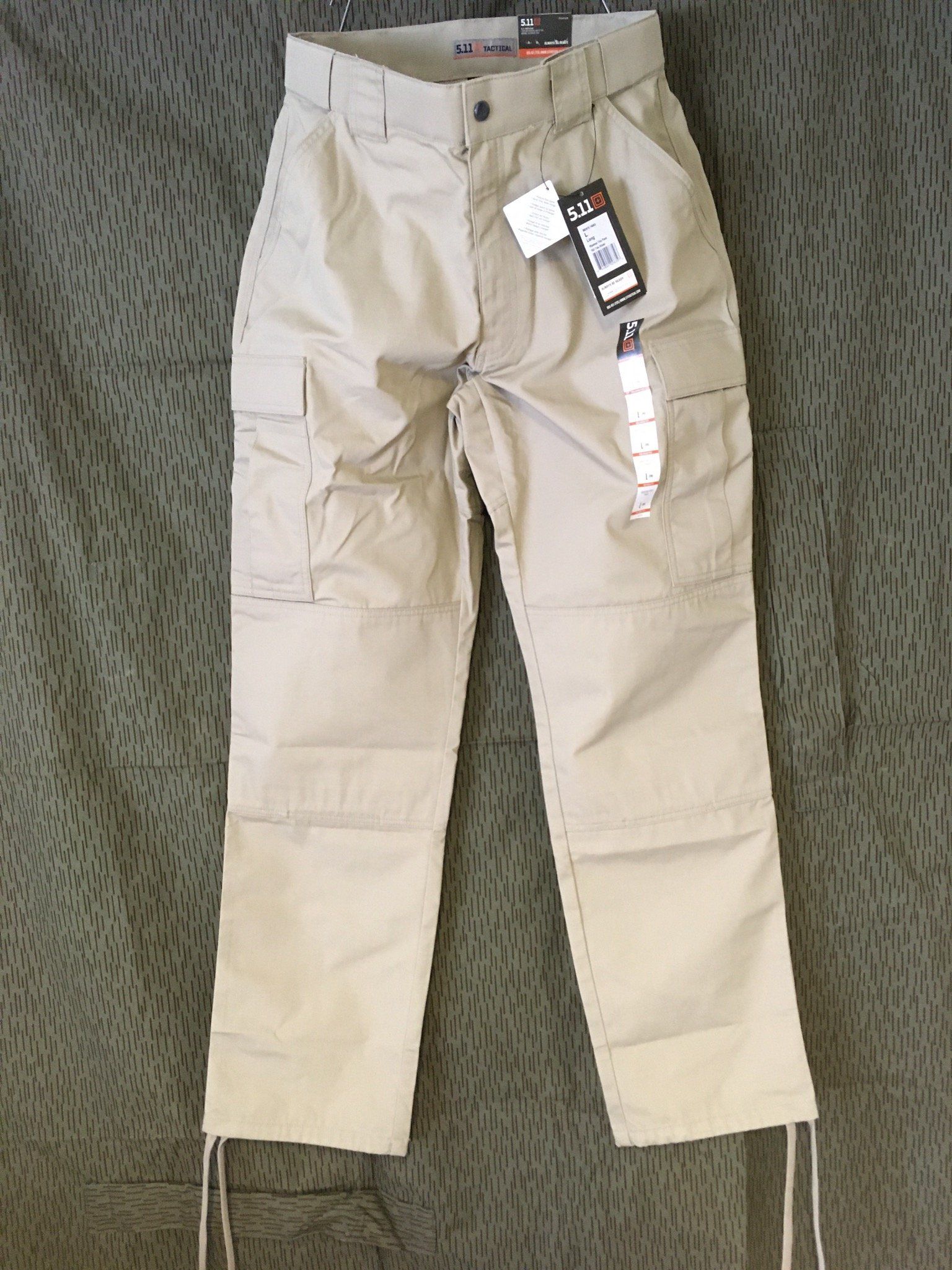 Khakifarbene Cargohose mit Etikett, flach auf einer strukturierten grünen Oberfläche liegend.