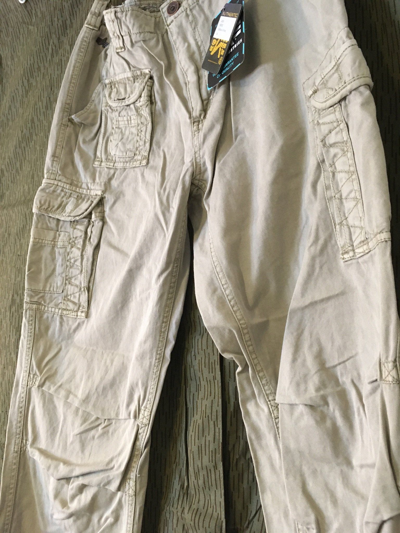Khakifarbene Cargohose mit mehreren Taschen und einem Etikett, die vor einem hölzernen Hintergrund hängt.