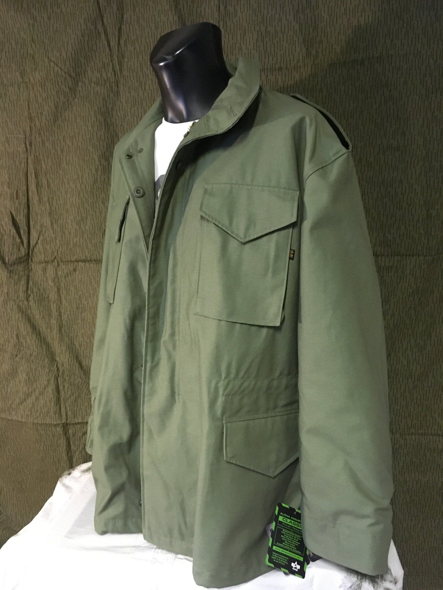 Grüne Jacke im Militärstil auf einer schwarzen Schaufensterpuppe vor einem grün gemusterten Hintergrund.